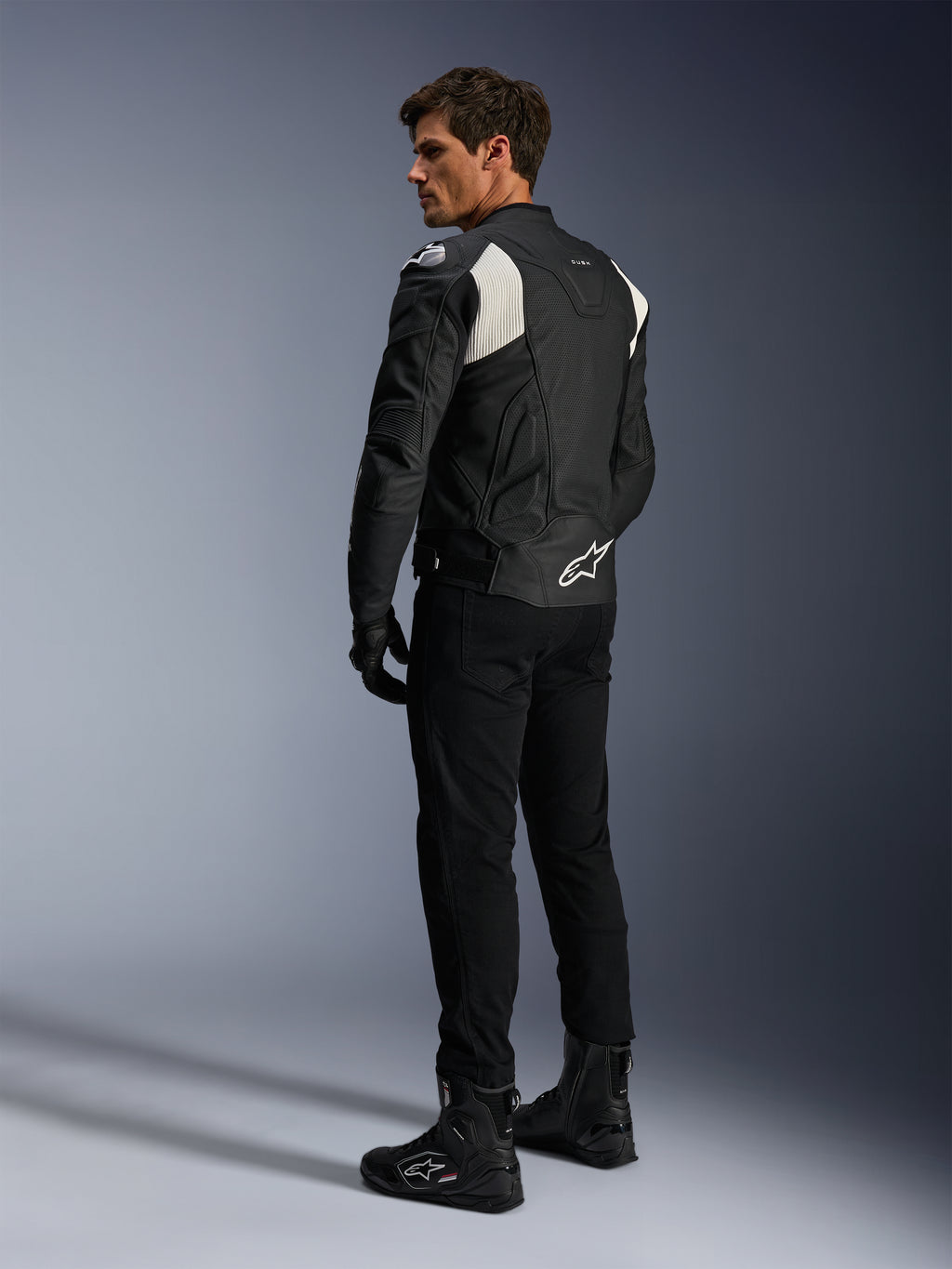 Alpinestars Dusk Airflow læderjakke, sort og hvid, set bagfra af model iført den perforerede læderjakke med matchende sorte bukser og støvler, hvide harmonika-strækpaneler og markant logo på lænden, sporty motorcykeludstyr