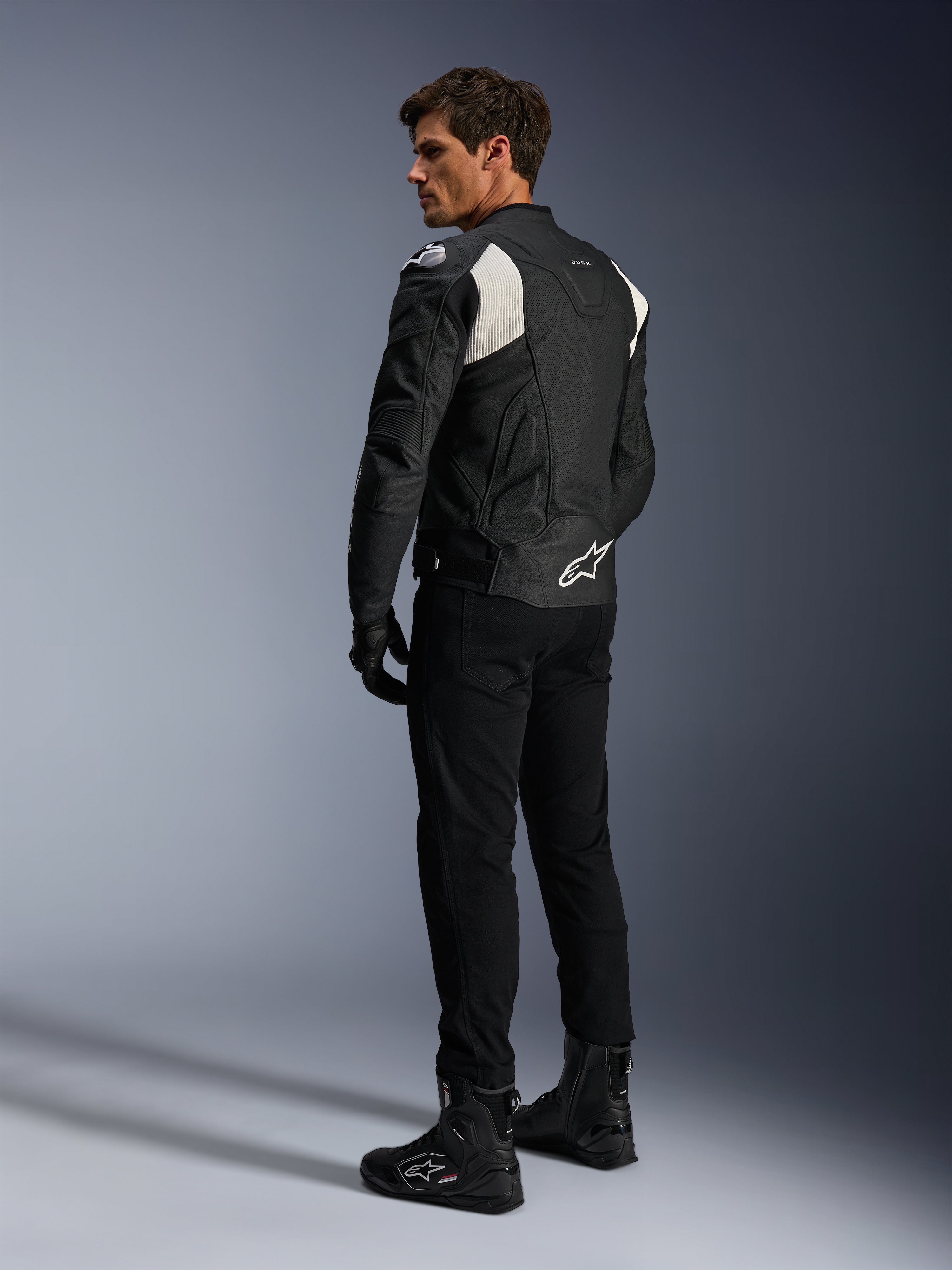 Alpinestars Dusk Airflow læderjakke, sort og hvid, set bagfra af model iført den perforerede læderjakke med matchende sorte bukser og støvler, hvide harmonika-strækpaneler og markant logo på lænden, sporty motorcykeludstyr