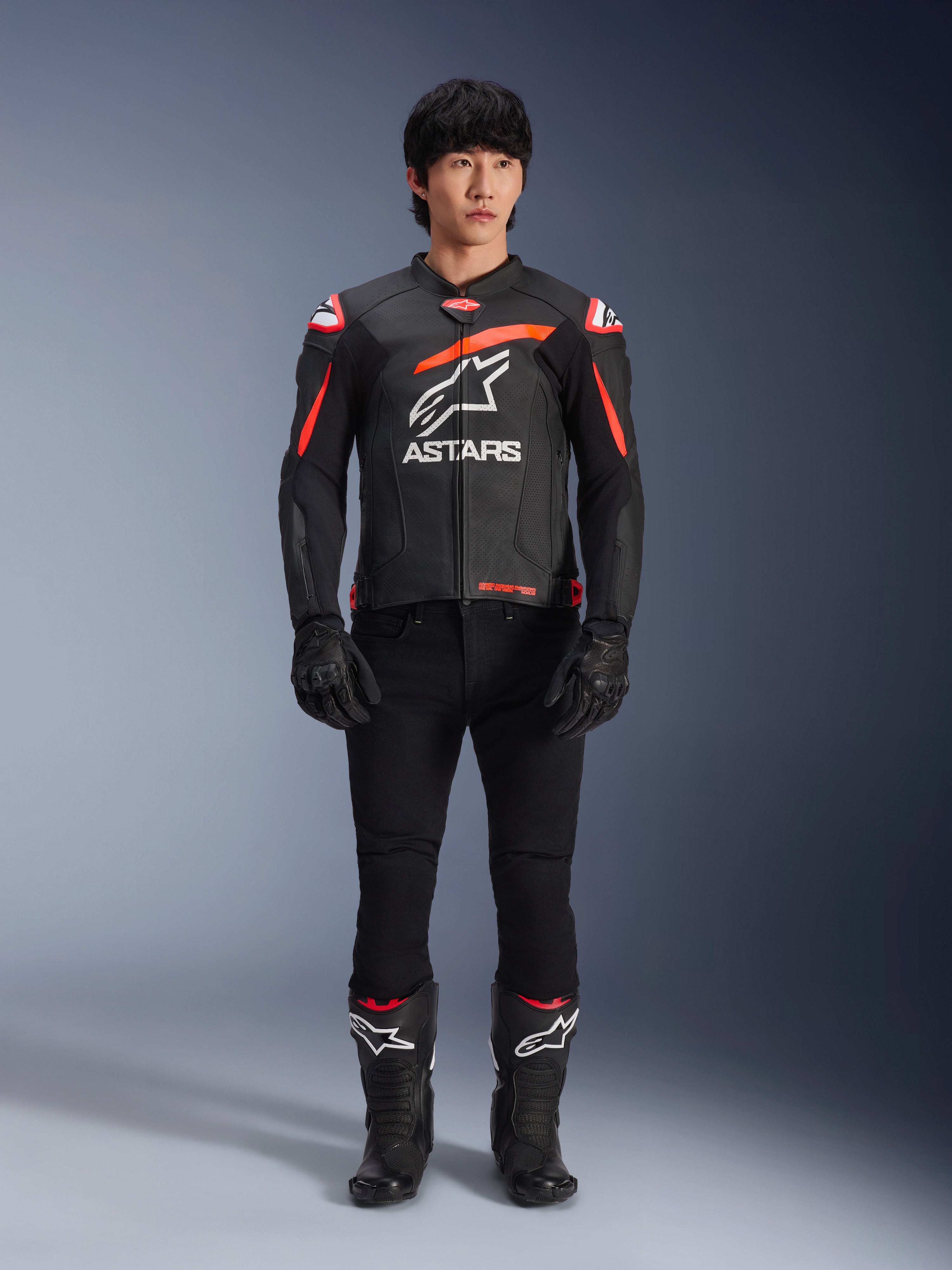 Mandlig model iført Alpinestars GP Plus R V4 Airflow læderjakke, sort med rød fluo og hvide detaljer, stort hvidt Astars-logo på brystet, kombineret med sorte tekniske bukser, handsker og støvler, højtydende motorcykeludstyr i et studiomiljø