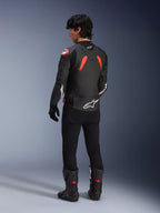 Set bagfra af en model iført Alpinestars GP Plus R V4 Airflow læderjakke, sort med rød fluo og hvide detaljer, båret med sorte bukser og sorte motorcykelstøvler, med fremtrædende hvide logoer på ryggen og ærmerne, læderkonstruktion i sportsstil