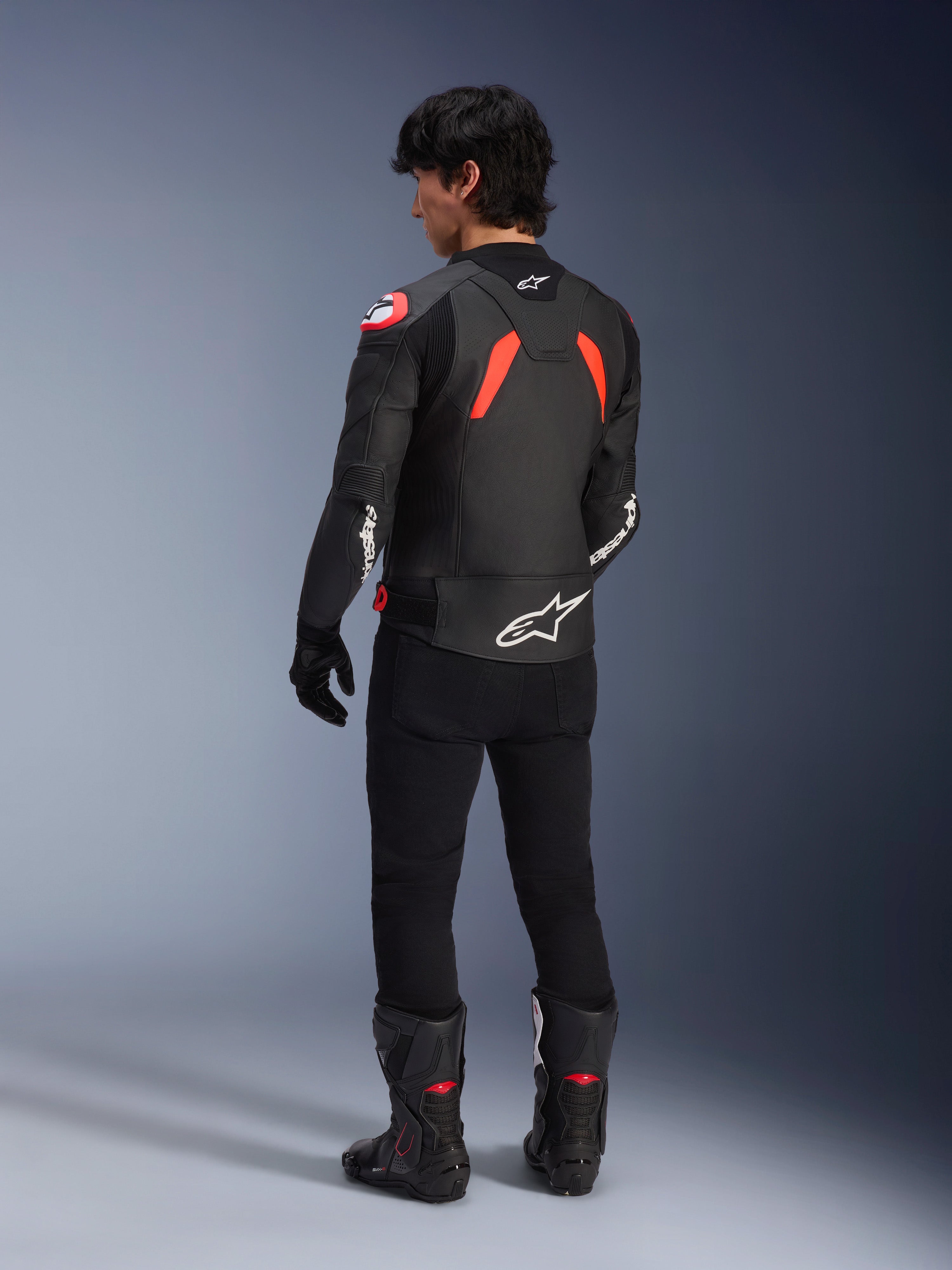 Set bagfra af en model iført Alpinestars GP Plus R V4 Airflow læderjakke, sort med rød fluo og hvide detaljer, båret med sorte bukser og sorte motorcykelstøvler, med fremtrædende hvide logoer på ryggen og ærmerne, læderkonstruktion i sportsstil
