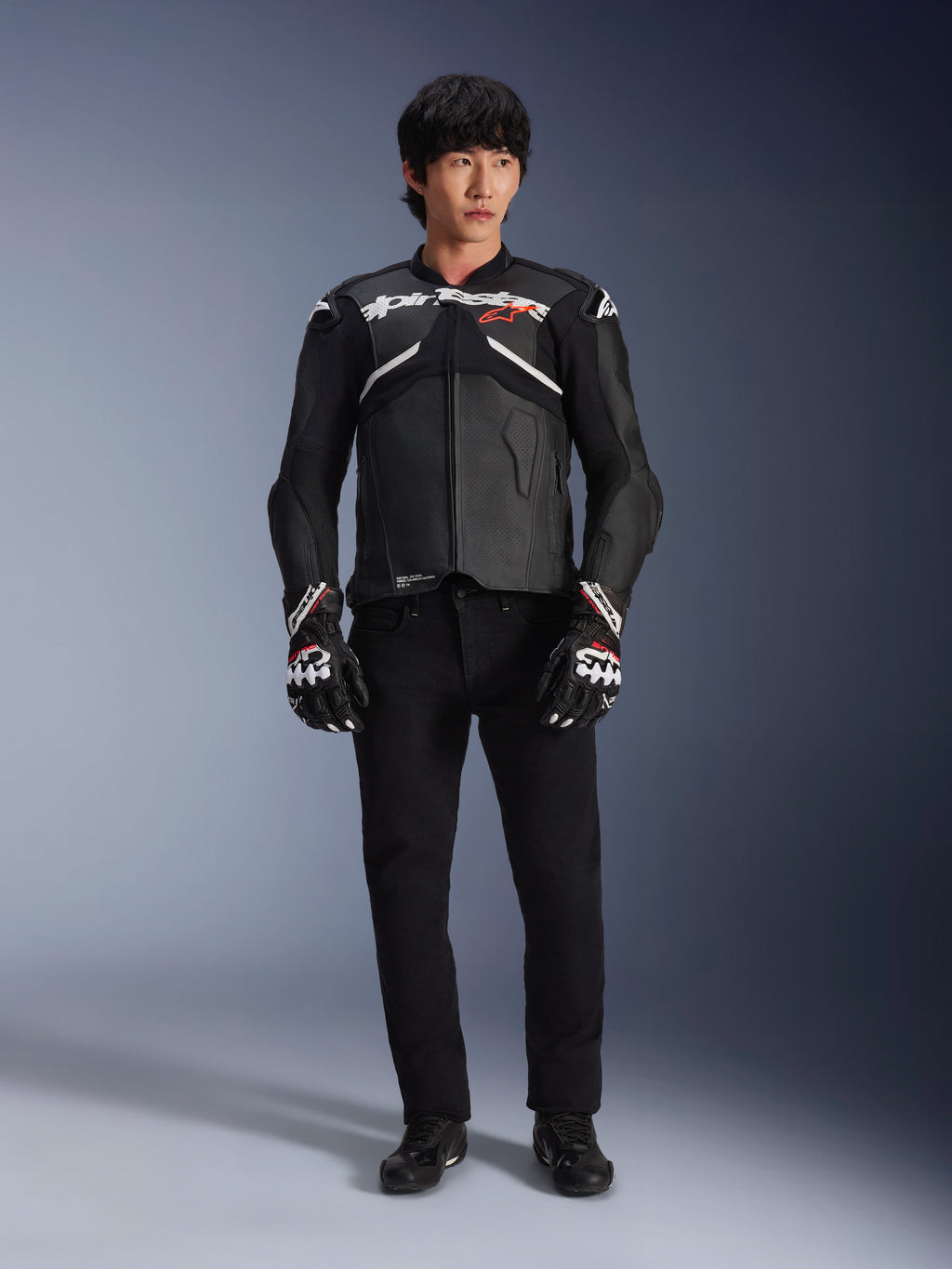Model iført Alpinestars Atem V5 læderjakke i sort og hvid, med hvidt Alpinestars-logo og et rødt stjernelogo på brystet, kombineret med sorte bukser og sort-hvide racinghandsker, strømlinet performance motorcykeludstyr