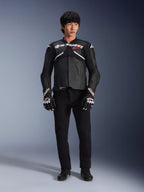 Model iført Alpinestars Atem V5 læderjakke i sort og hvid, med hvidt Alpinestars-logo og et rødt stjernelogo på brystet, kombineret med sorte bukser og sort-hvide racinghandsker, strømlinet performance motorcykeludstyr