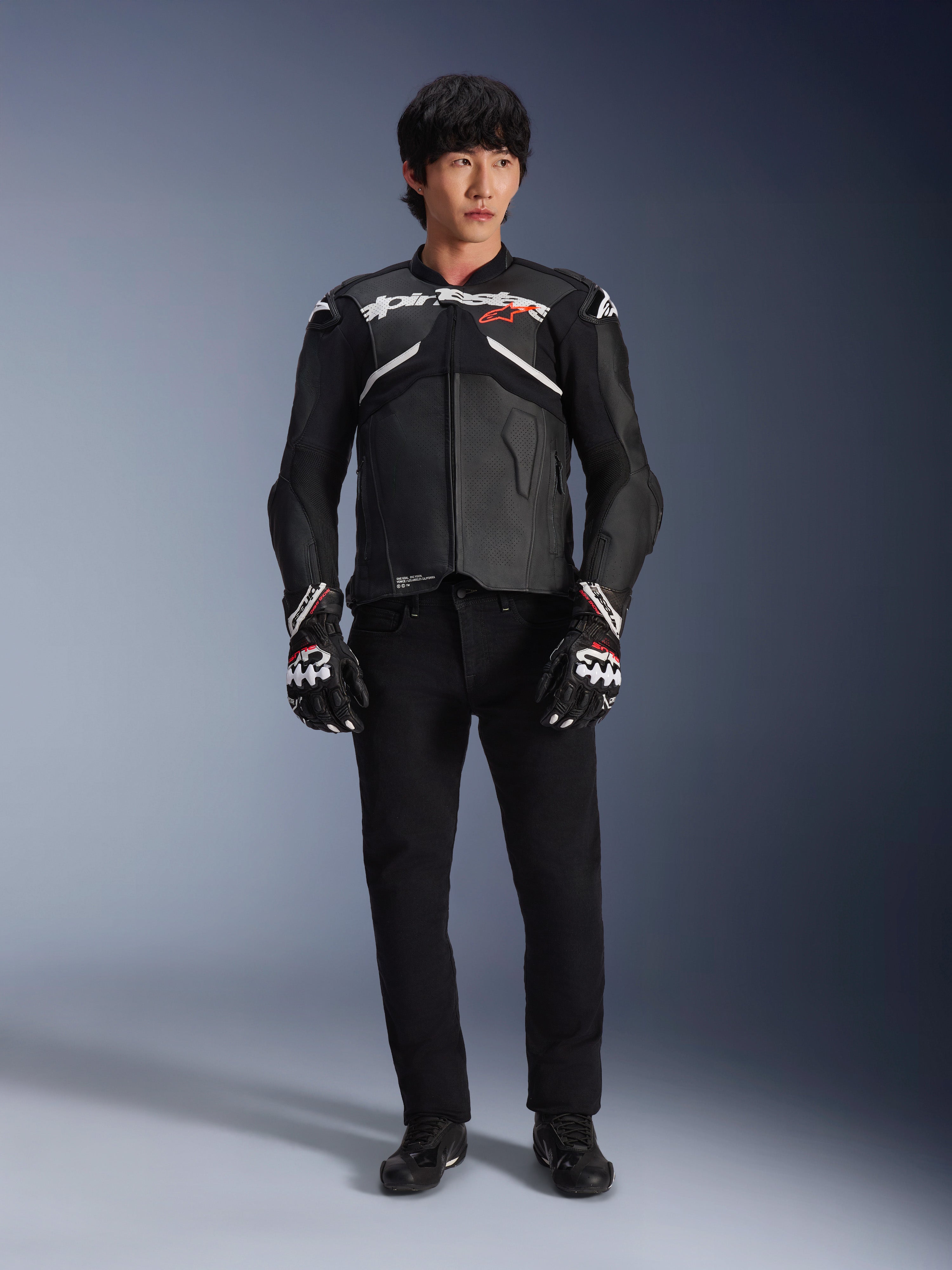 Model iført Alpinestars Atem V5 læderjakke i sort og hvid, med hvidt Alpinestars-logo og et rødt stjernelogo på brystet, kombineret med sorte bukser og sort-hvide racinghandsker, strømlinet performance motorcykeludstyr