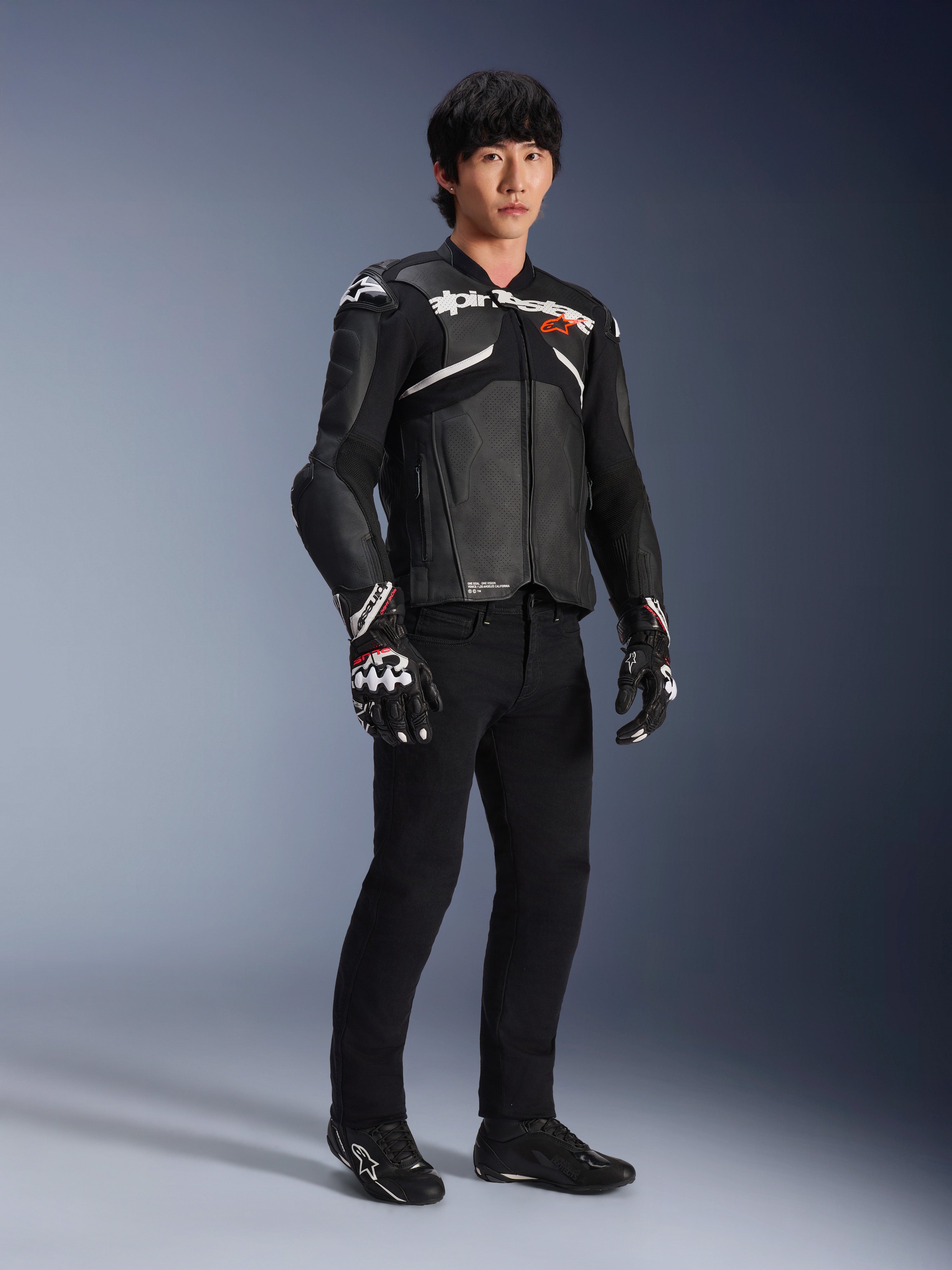 Alpinestars Atem V5 læderjakke, motorcykel racerjakke, sort og hvid, båret med sorte bukser og matchende handsker, med hvid Alpinestars-branding over brystet og beskyttende skulderbeskyttelse.
