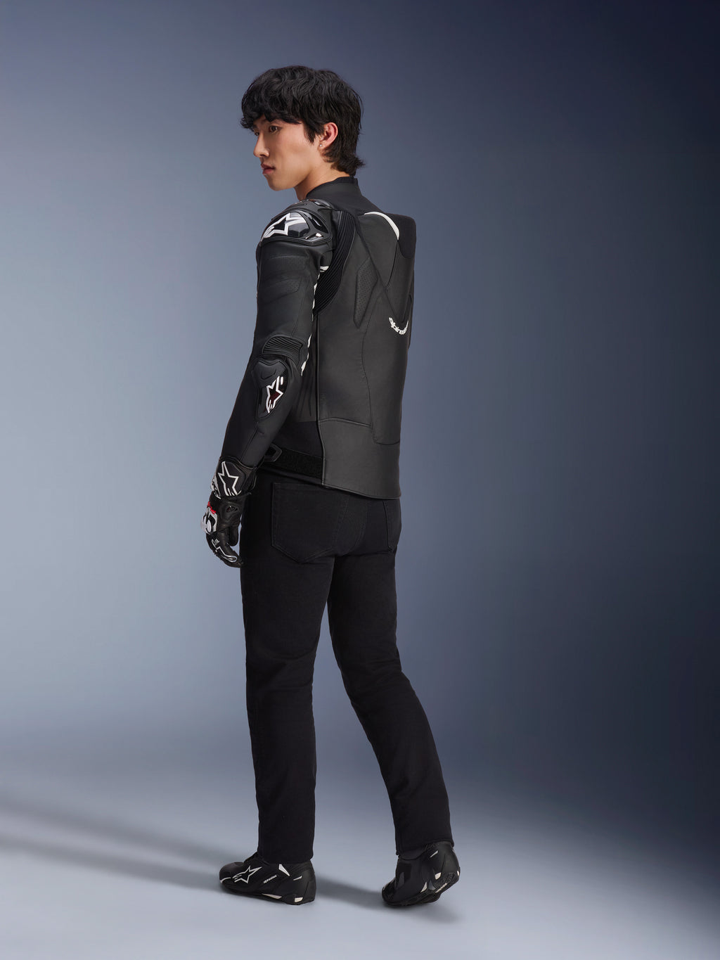 Mandlig model iført Alpinestars Atem V5-læderjakken i sort og hvid, trekvart visning bagfra, der viser den aerodynamiske hastighedspukkel og DFS RR-beskyttere, kombineret med sorte bukser, racerhandsker og støvler, professionelt motorcykel-racerudstyr