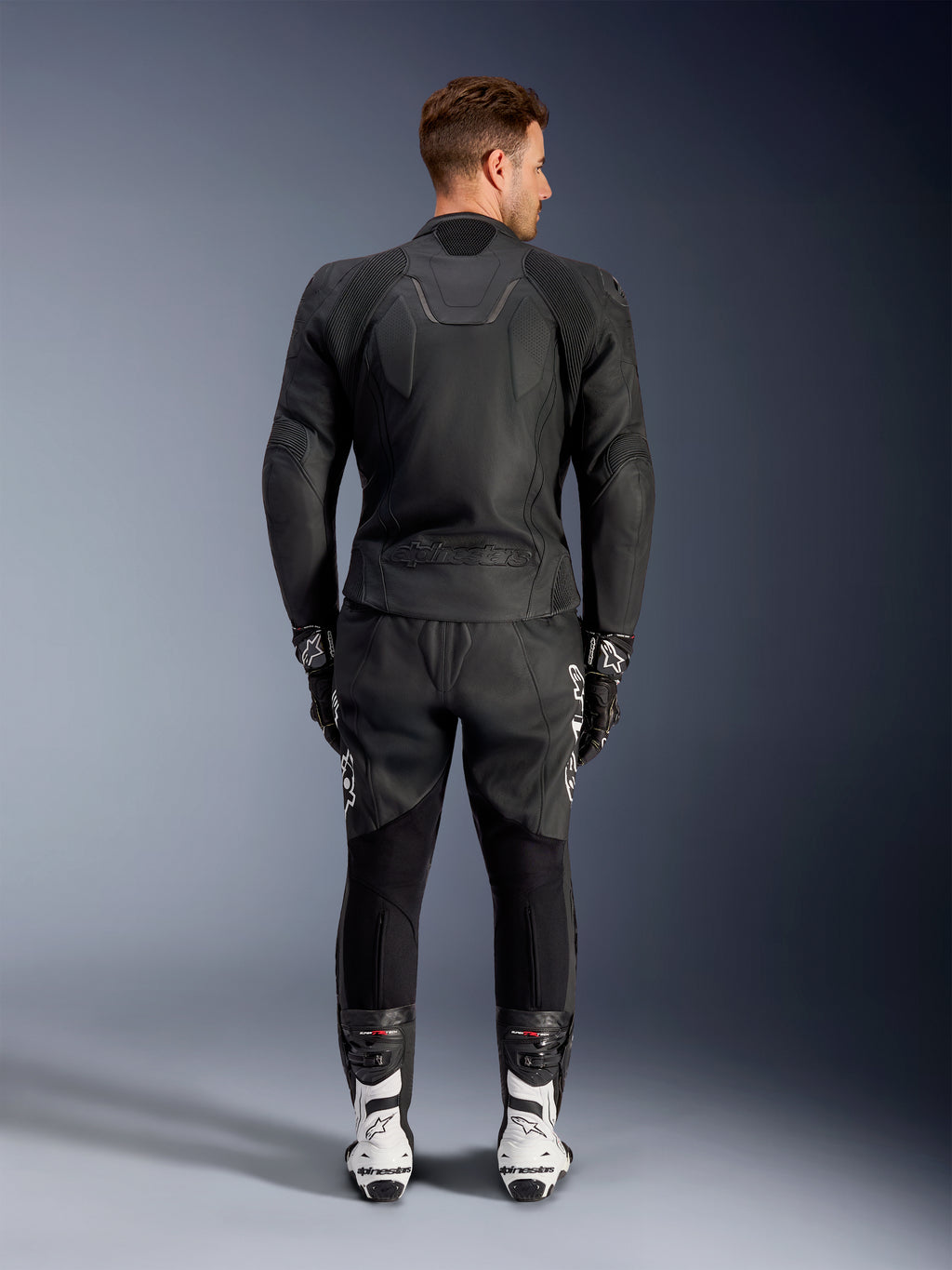 Bagfra-visning af en mand iført Alpinestars Caliber læderjakke i sort, med præget logo og perforerede læderpaneler, kombineret med sorte motorcykelbukser i læder, sorte handsker og hvide racingstøvler.