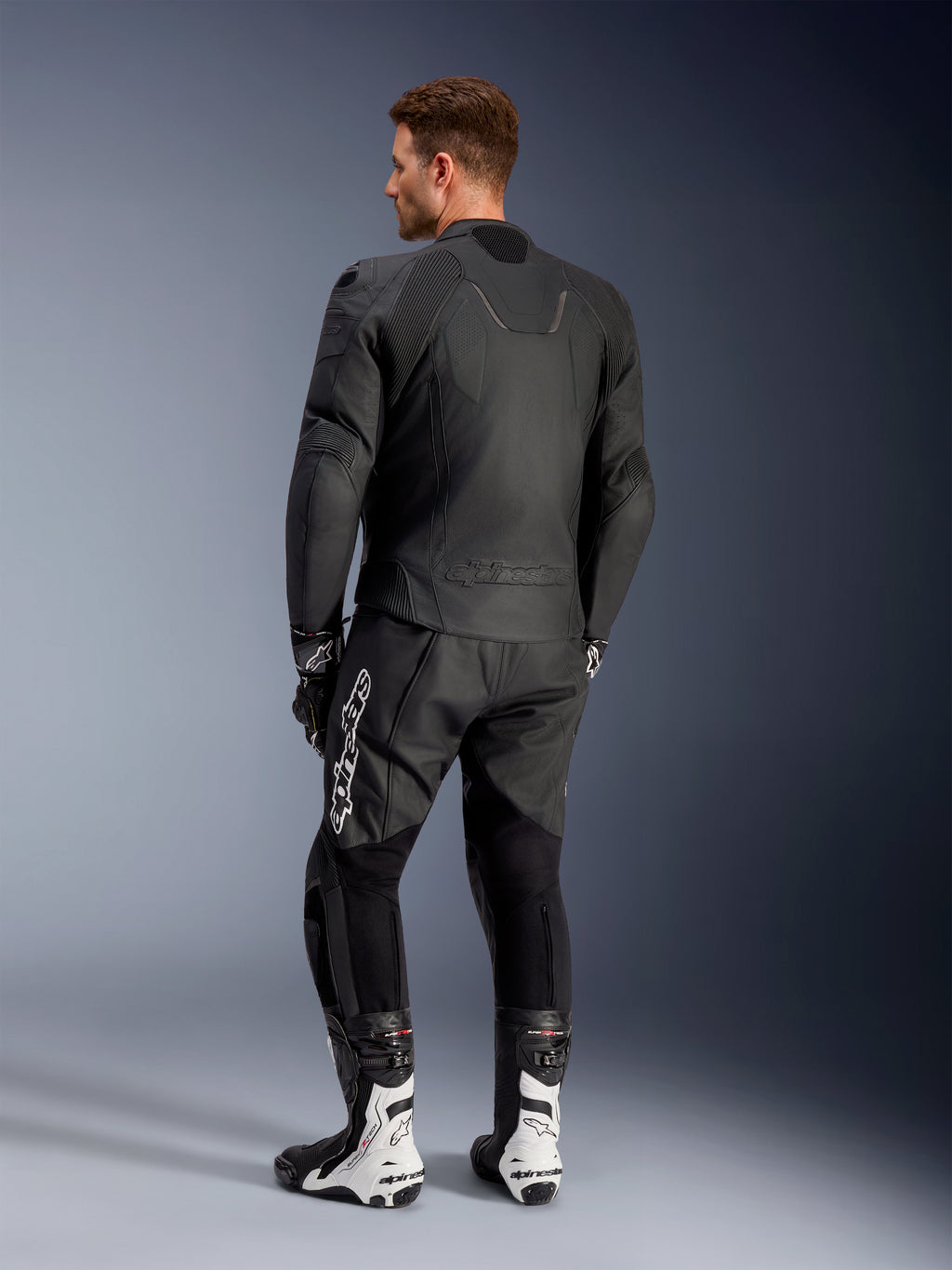 Alpinestars Caliber læderjakke, sort motorcykeljakke til vejbrug, set bagfra, med perforeret læder og akkordeon-strækindsatser ved taljen og skuldrene, præget logo, båret med sorte læderbukser og hvide racingstøvler