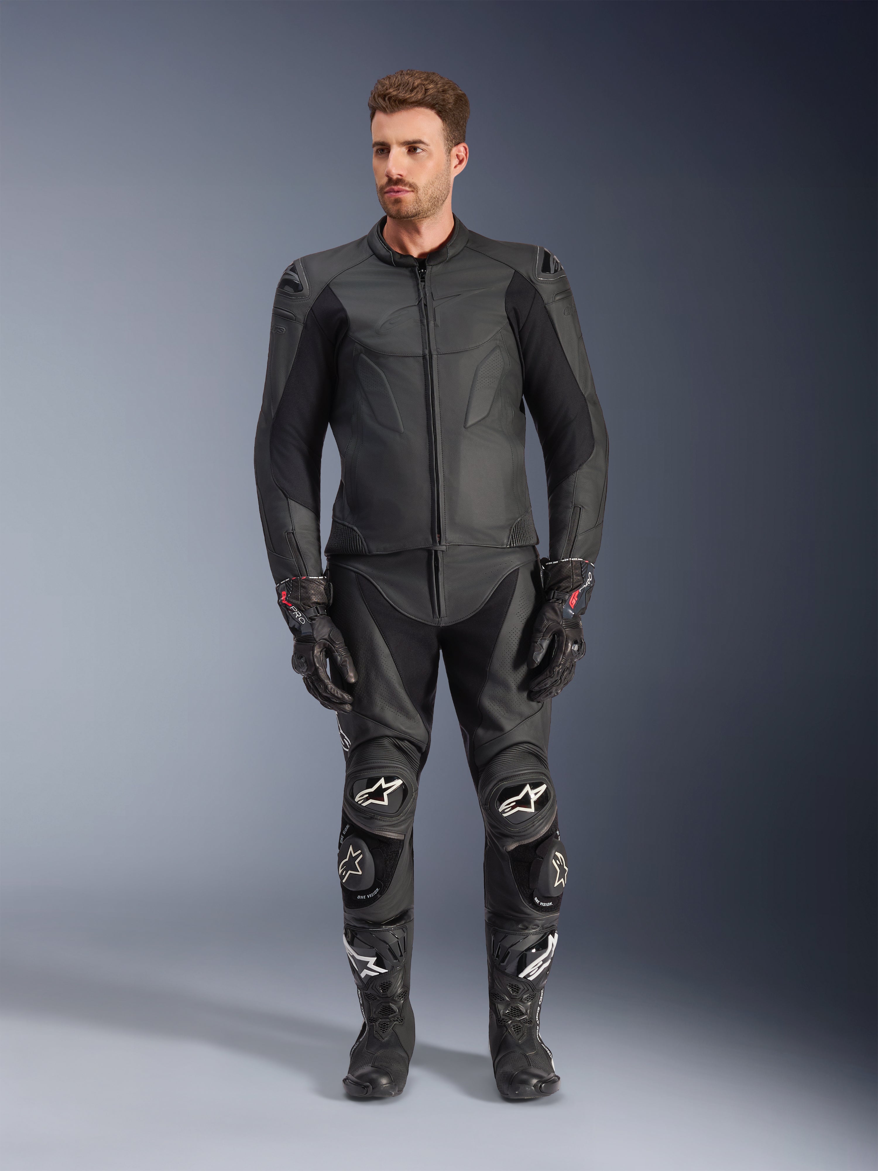 Alpinestars Caliber læderjakke, sort motorcykeljakke, båret med matchende racerbukser, handsker og støvler, med et strømlinet design med tonale logoer, strækpaneler på ærmerne og beskyttende skulderslidere, professionel studieopsætning