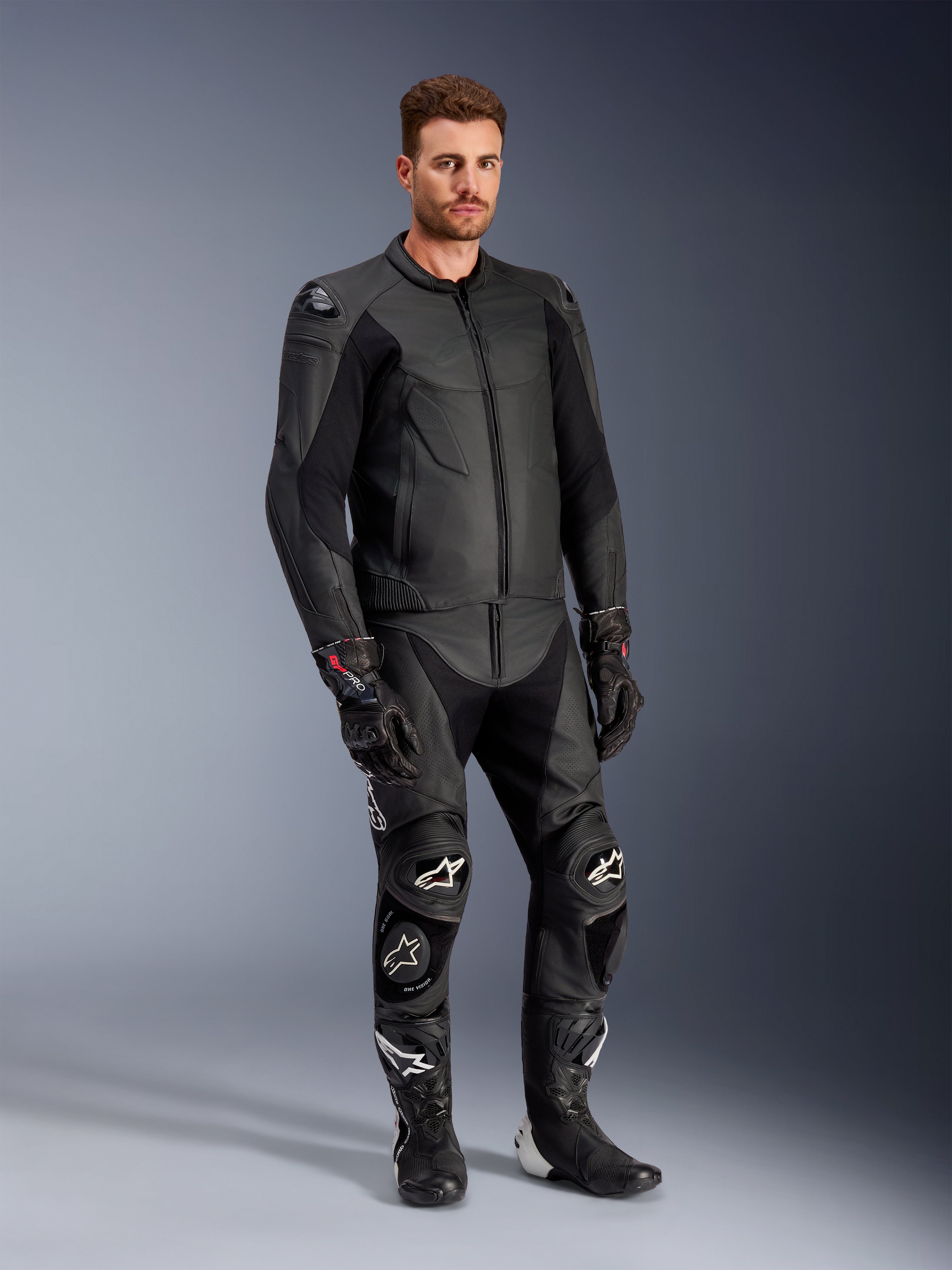 Mandlig model iført Alpinestars Caliber læderjakke i sort, kombineret med matchende sorte lædermotorcykelbukser, racinghandsker og støvler, som viser et komplet sportsligt motorcykeloutfit til vej.