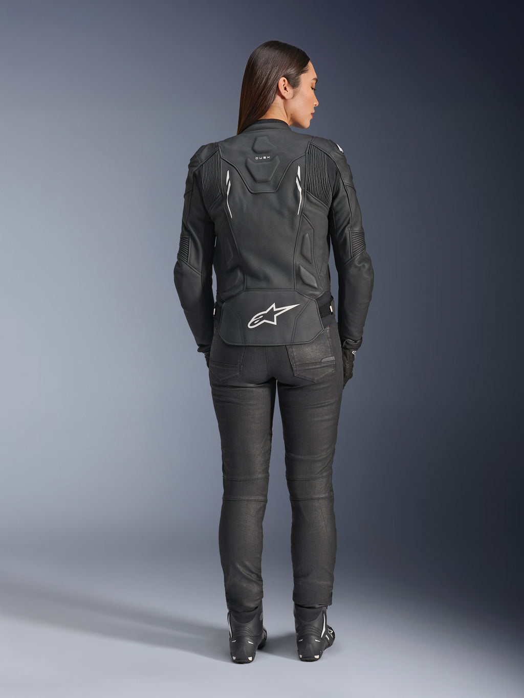 Alpinestars Stella Dusk læderjakke til kvinder, sort, set bagfra med hvidt Alpinestars-logo på den nederste del af ryggen og 'DUSK'-branding øverst, båret med sorte kørebukser og støvler, hvilket viser den konturerede kvindespecifikke pasform og beskyttende læderkonstruktion.