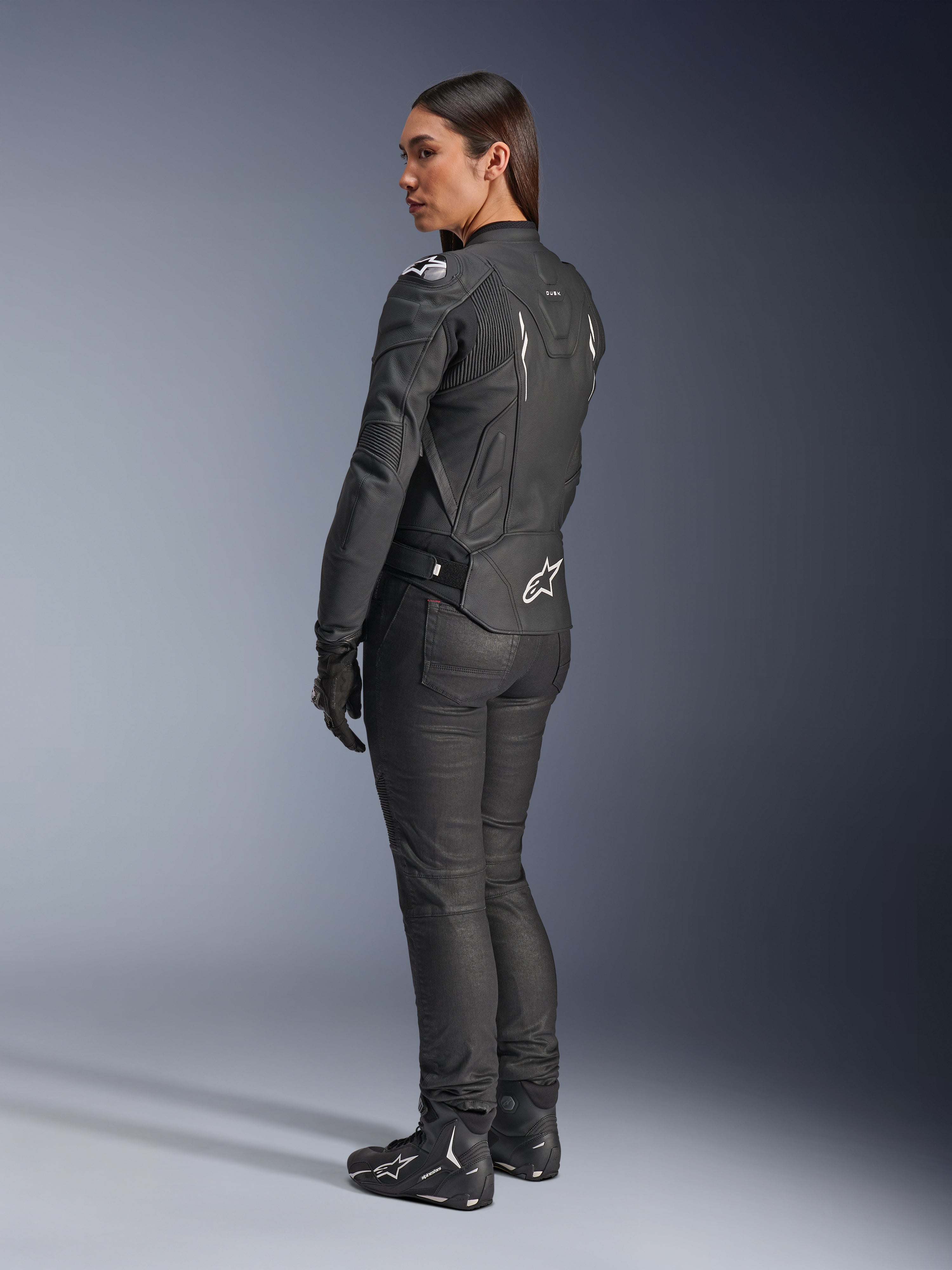 Alpinestars Stella Dusk læderjakke, motorcykeljakke til kvinder, sort og hvid, set bagfra med hvidt Alpinestars-logo på den nederste del af ryggen og skuldrene, båret med sorte kørebukser og støvler, sportsfokuseret ergonomisk pasform til landevejskørsel