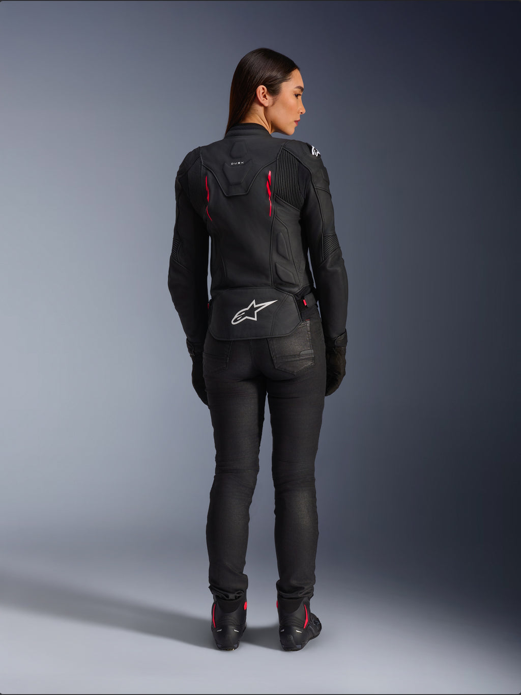 Alpinestars Stella Dusk læderjakke, set bagfra, sort læder med Diva Pink og hvide detaljer, hvidt Alpinestars-logo nederst på ryggen og lyserøde ventilationsdetaljer, båret med sorte kørebukser og støvler, premium sportsmotorcykeludstyr til kvinder
