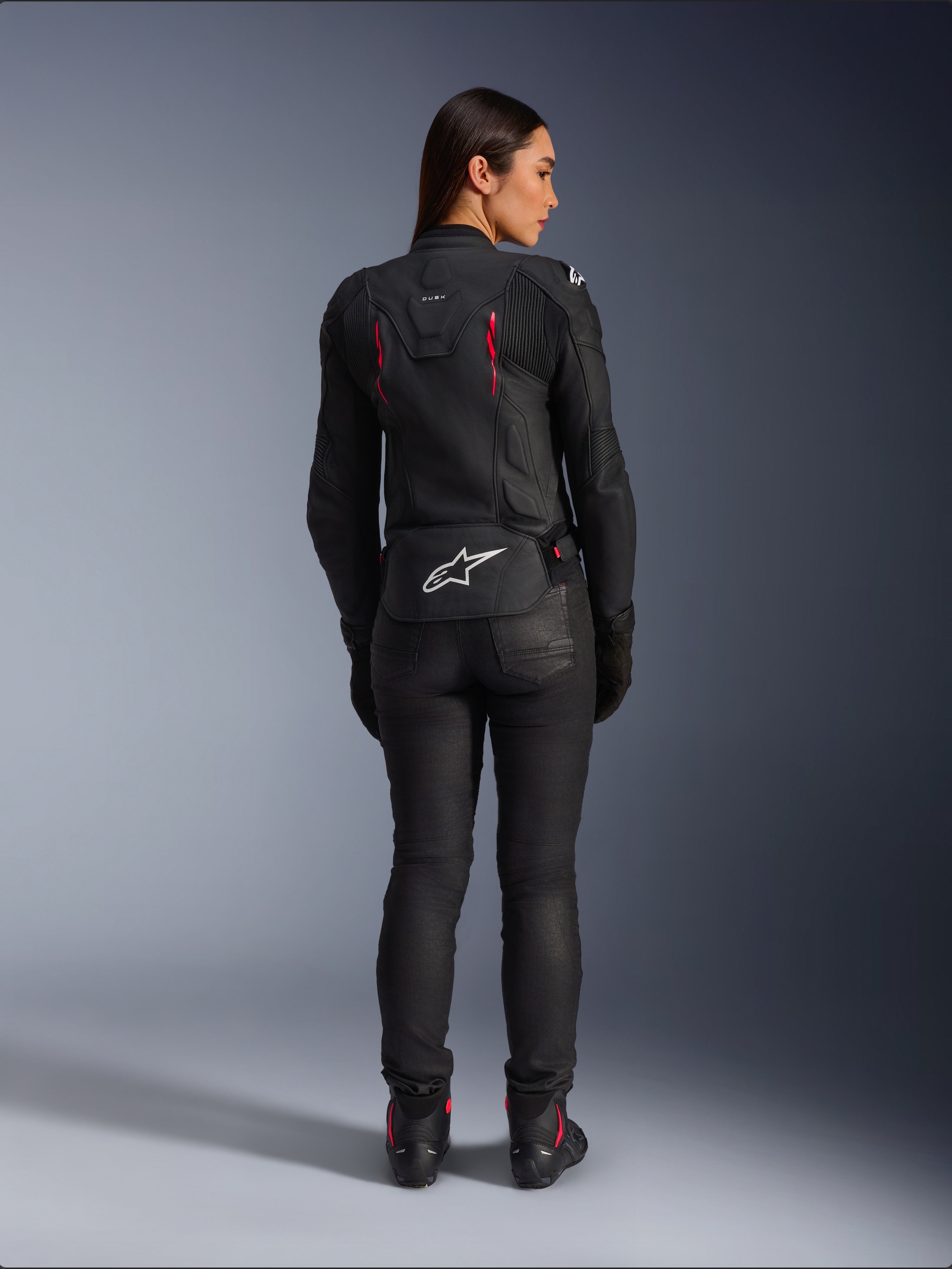 Alpinestars Stella Dusk læderjakke, set bagfra, sort læder med Diva Pink og hvide detaljer, hvidt Alpinestars-logo nederst på ryggen og lyserøde ventilationsdetaljer, båret med sorte kørebukser og støvler, premium sportsmotorcykeludstyr til kvinder