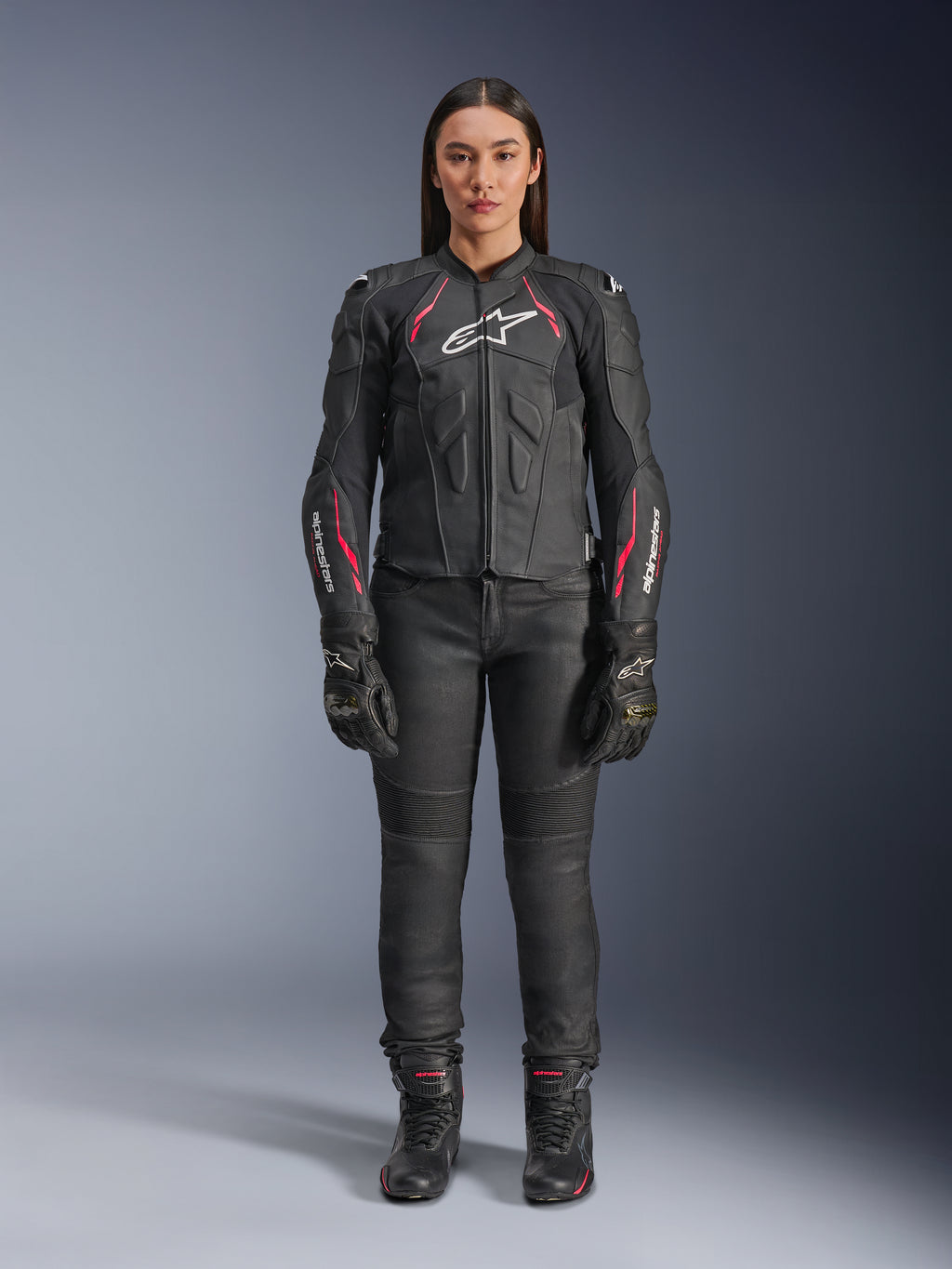 En kvinde iført Alpinestars Stella Dusk læderjakke i Black Diva Pink White, med hvidt logo på brystet og lyserøde detaljer på skuldre og ærmer, kombineret med sorte motorcykelbukser, handsker og støvler.