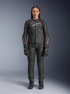 En kvinde iført Alpinestars Stella Dusk læderjakke i Black Diva Pink White, med hvidt logo på brystet og lyserøde detaljer på skuldre og ærmer, kombineret med sorte motorcykelbukser, handsker og støvler.