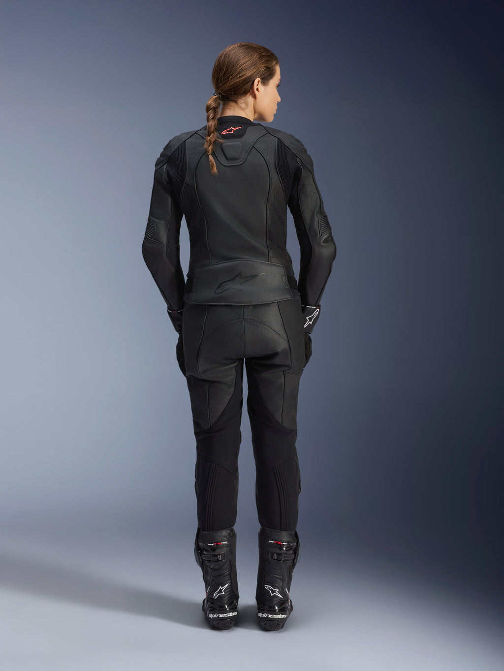 Alpinestars Stella GP Plus V4 læderjakke til kvinder, Black Black, fuldkropsvisning bagfra af en kvindelig rytter iført matchende læderbukser og støvler, strømlinet minimalistisk design med et lille rødt logo i nakken, skræddersyet sports-silhuet til kvinder