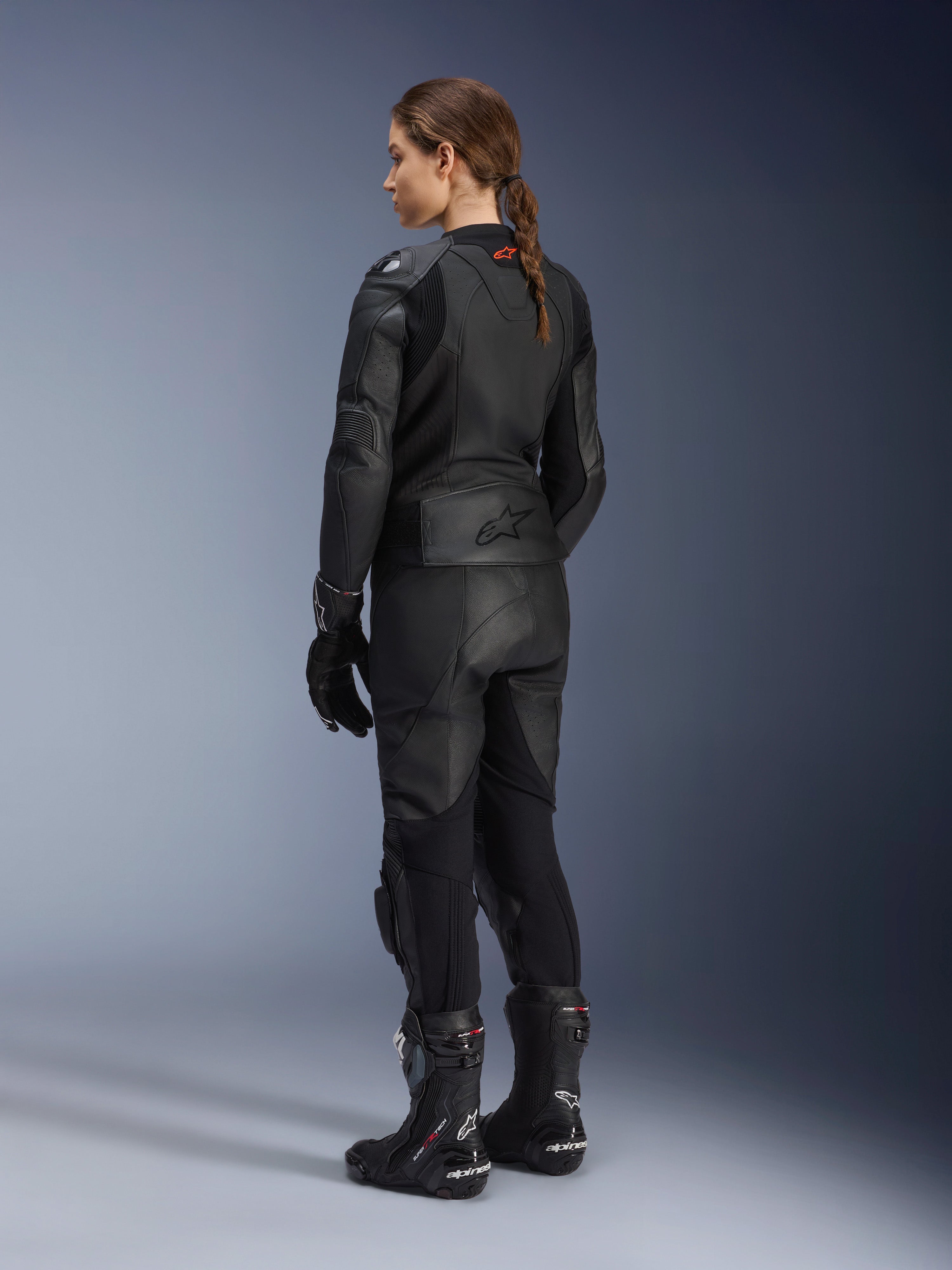 Alpinestars Stella GP Plus V4 læderjakke til kvinder, sort, fuld visning bagfra, kombineret med sorte motorcykelbukser i læder, racerhandsker og støvler, med præget Alpinestars-logo på lænden og strømlinet silhuet til sportskørsel