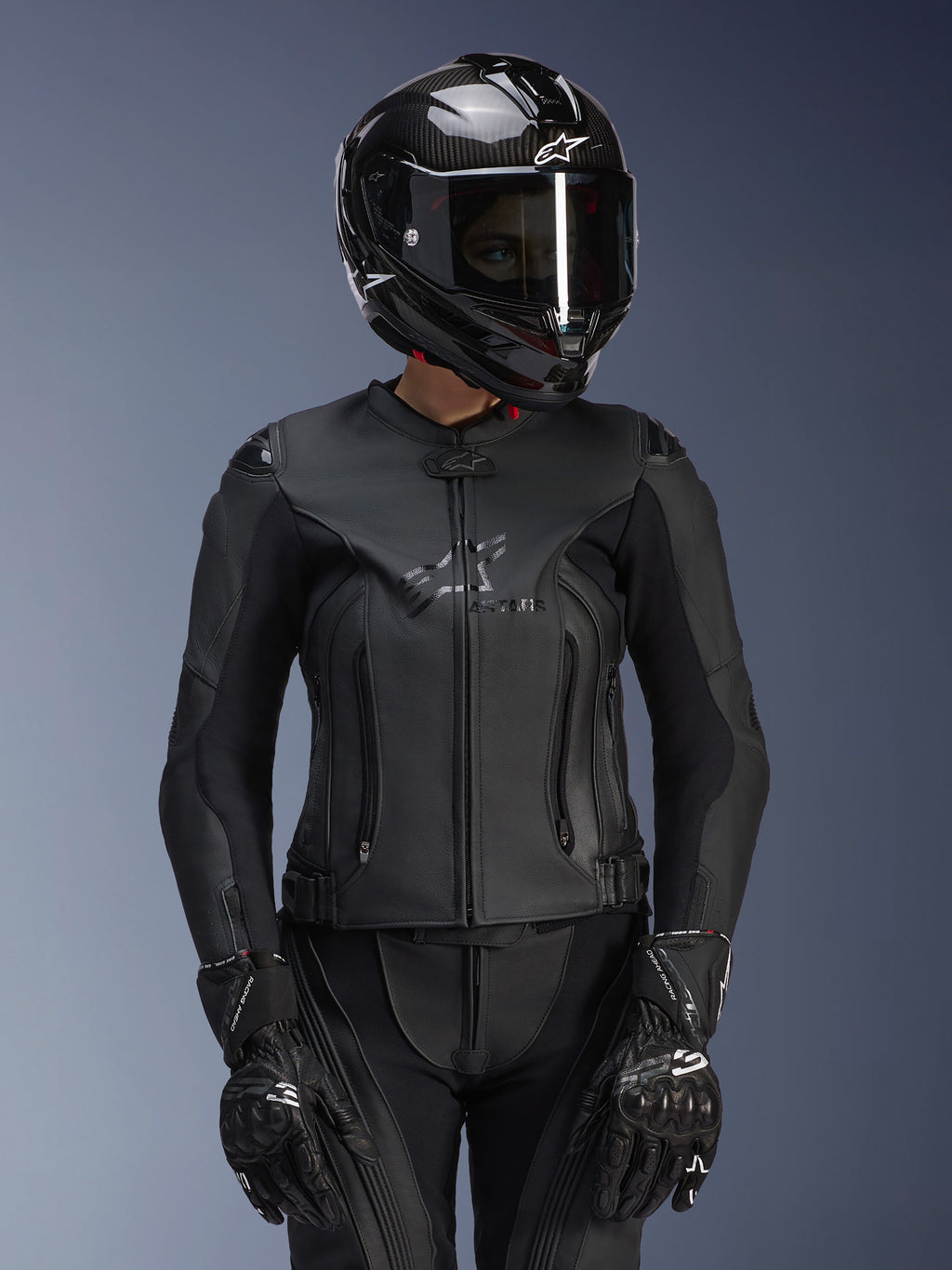 Alpinestars Stella GP Plus V4 læderjakke til kvinder i Black Black, vist med en sort kulfiberhjelm og racerhandsker, strømlinet læderkonstruktion med diskret branding på brystet, skræddersyet silhuet til kvindelige kørere, professionelt sportsmotorcykeludstyr
