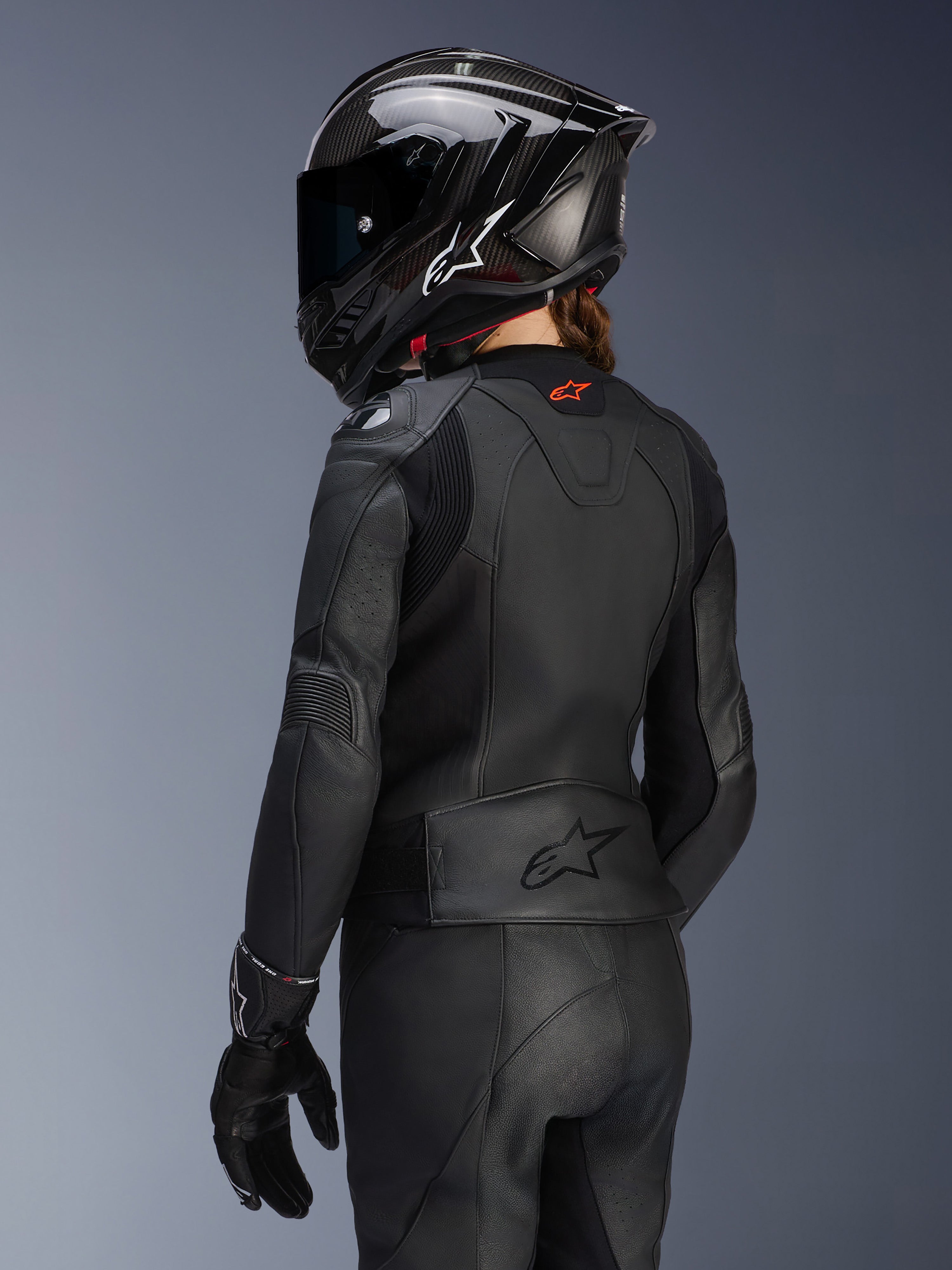 Kvindelig rytter set bagfra iført Alpinestars Stella GP Plus V4 læderjakke til kvinder i sort, matchende læderbukser og handsker samt en sort full-face hjelm i carbon-stil, der fremhæver jakkens aerodynamiske pasform og prægede logo på den nederste del af ryggen.