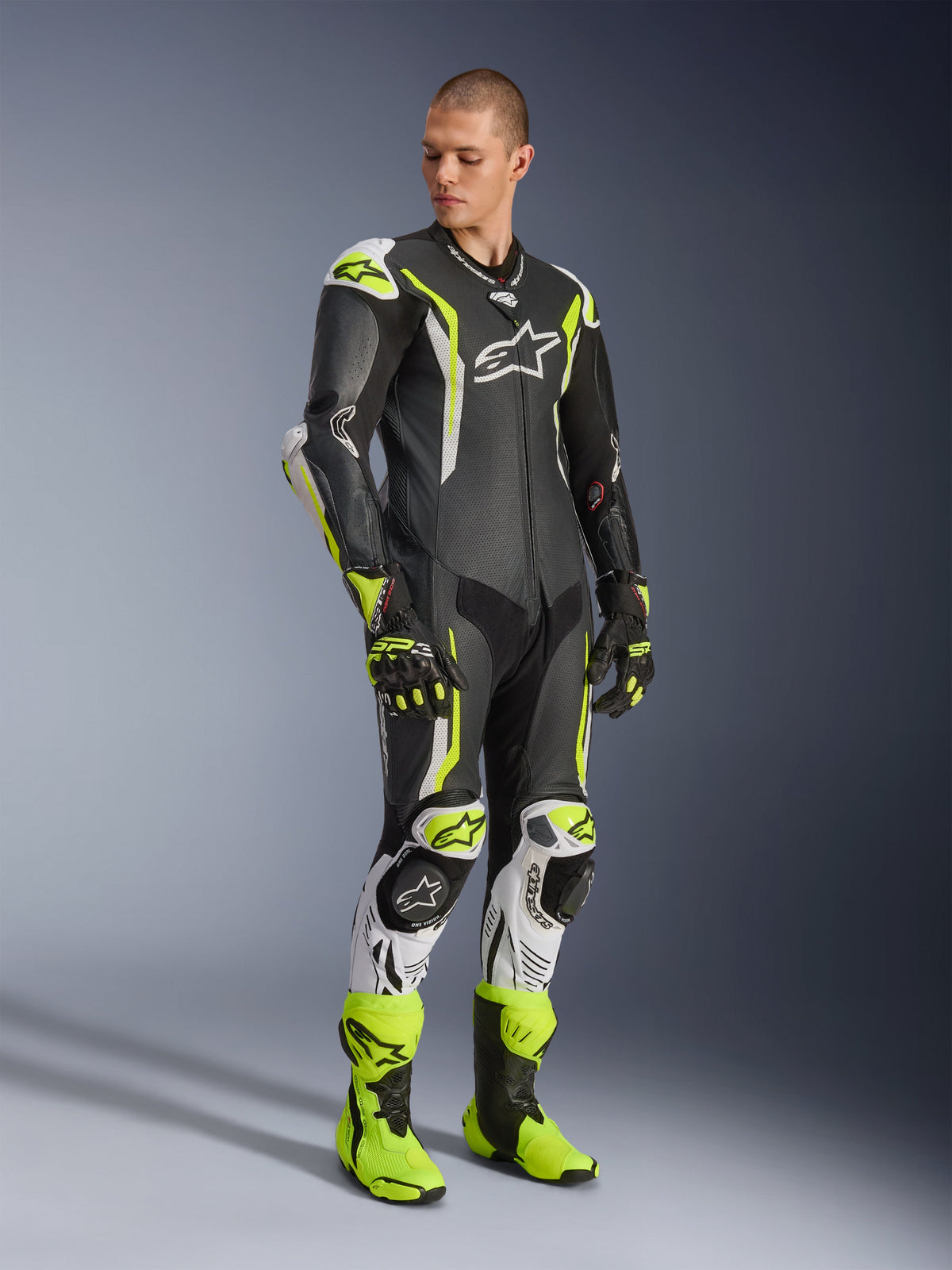 Alpinestars GP Tech V5 læderdragt i ét stykke, sort, hvid og gul fluo, båret af en model med matchende racinghandsker og støvler, professionelt motorcykel-racingudstyr med aerodynamisk ydeevne og beskyttelse
