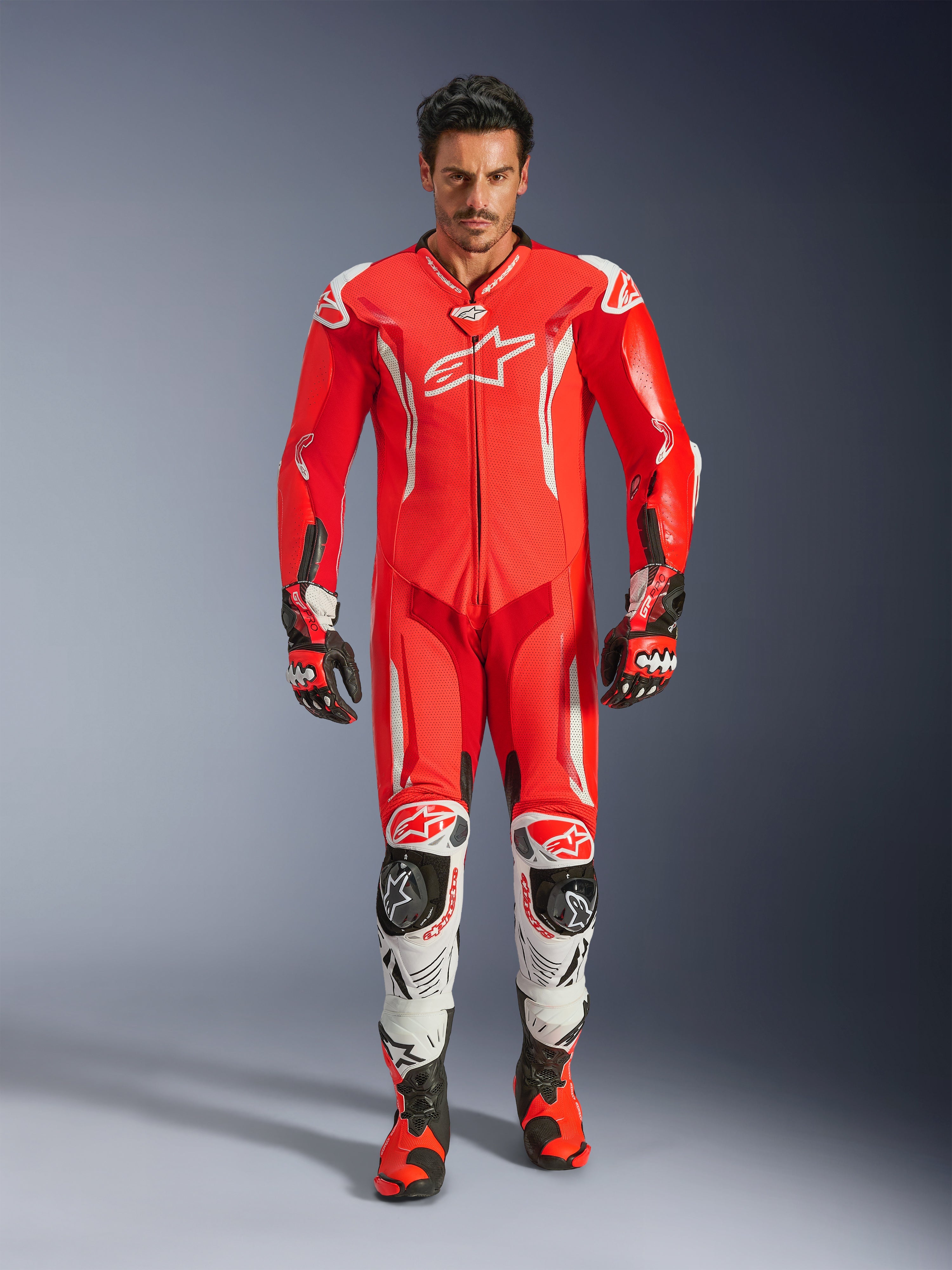 Alpinestars GP Tech V5 læderdragt i ét stykke, professionel racerdragt i Red Fluo og Bright Red med hvide detaljer, båret af en kører med matchende støvler og handsker, med en aerodynamisk hastighedspukkel, førsteklasses læderkonstruktion og avanceret beskyttelse til banekørsel.