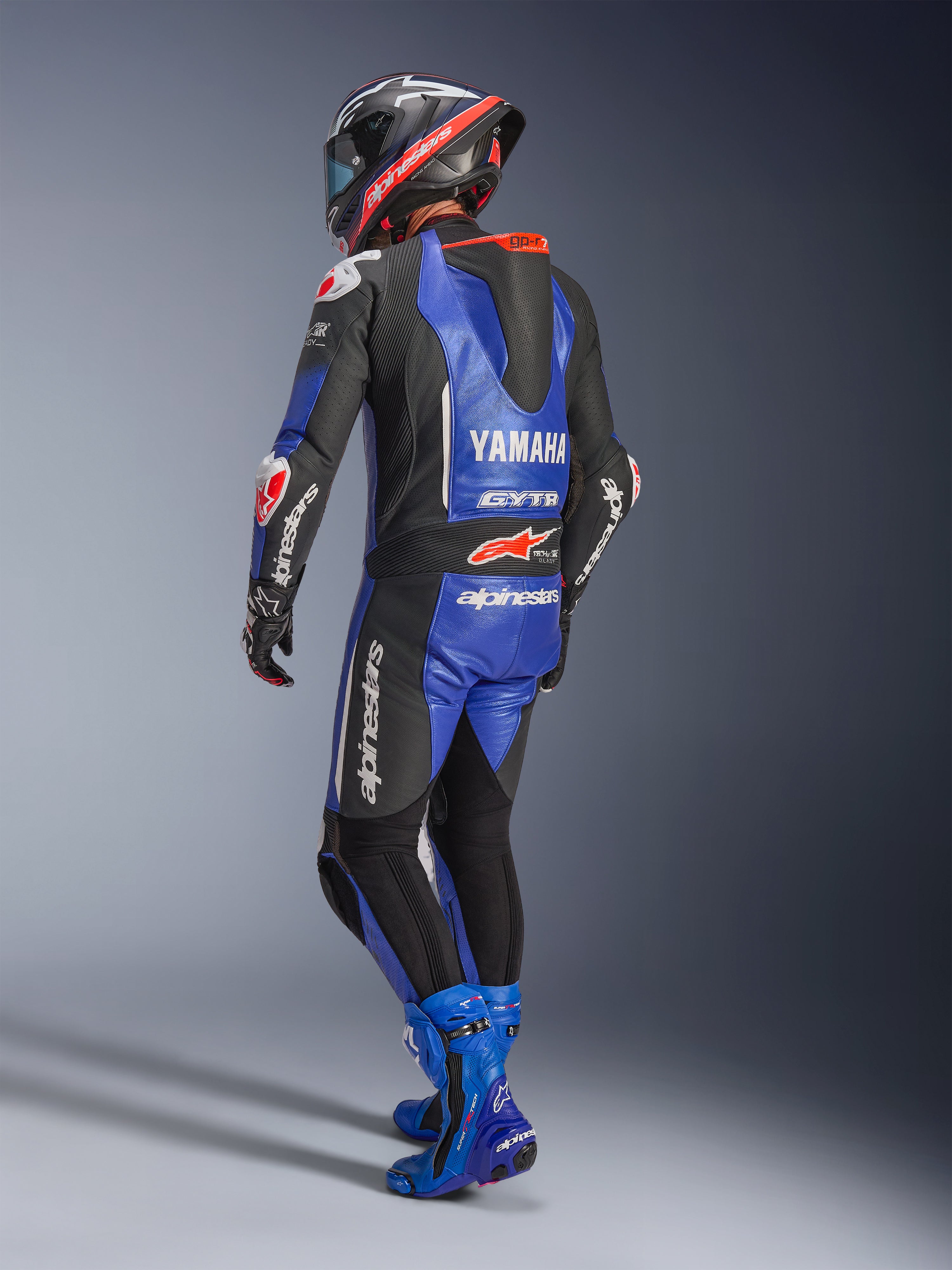 Alpinestars Yamaha GP-R7 læderdragt, metallisk blå, sort og hvid, set bagfra med aerodynamisk race-pukkel og Yamaha-branding, båret med matchende blå racerstøvler og hjelm, professionel performance-læderdragt til banekørsel