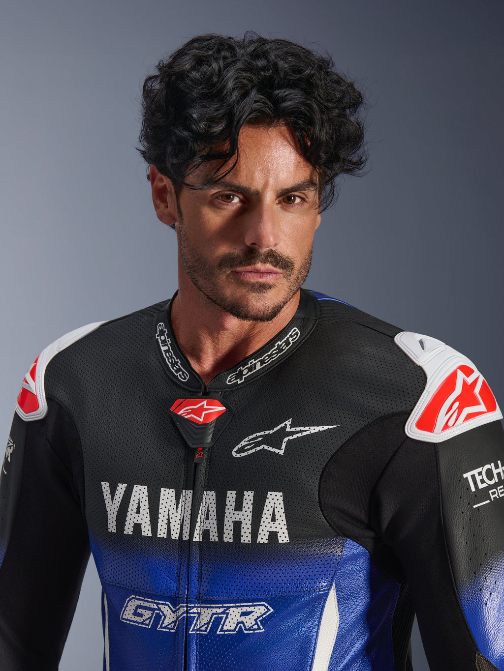 Nærbillede af en mand iført Alpinestars Yamaha GP-R7 1PC læderdragt, metallic blå og sort med hvid YAMAHA og GYTR branding, perforeret læder, hvide skulder-sliders med røde stjernelogoer, Tech-Air Ready, professionelt motorcykel-racerudstyr.