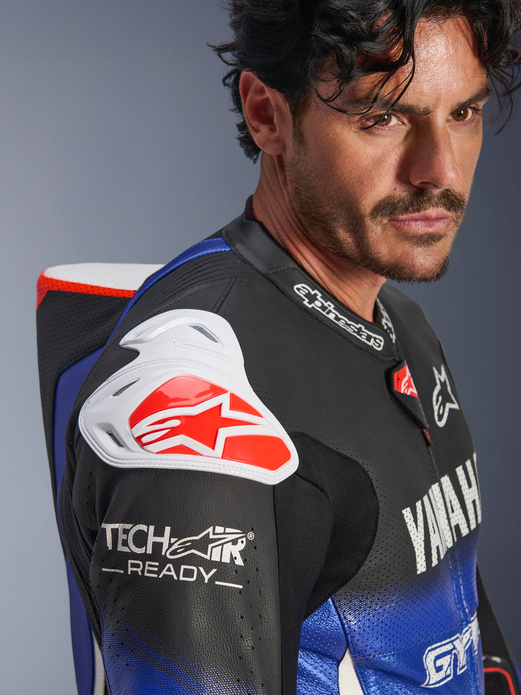 Yamaha GP-R7 læderdragt i ét stykke, metallic blå, sort, hvid og rød, nærbillede af skulderen og brystet, med hvid DFS-skulderbeskyttelse med et rødt logo, aerodynamisk pukkel og Tech-Air Ready-branding på det perforerede læderærme.