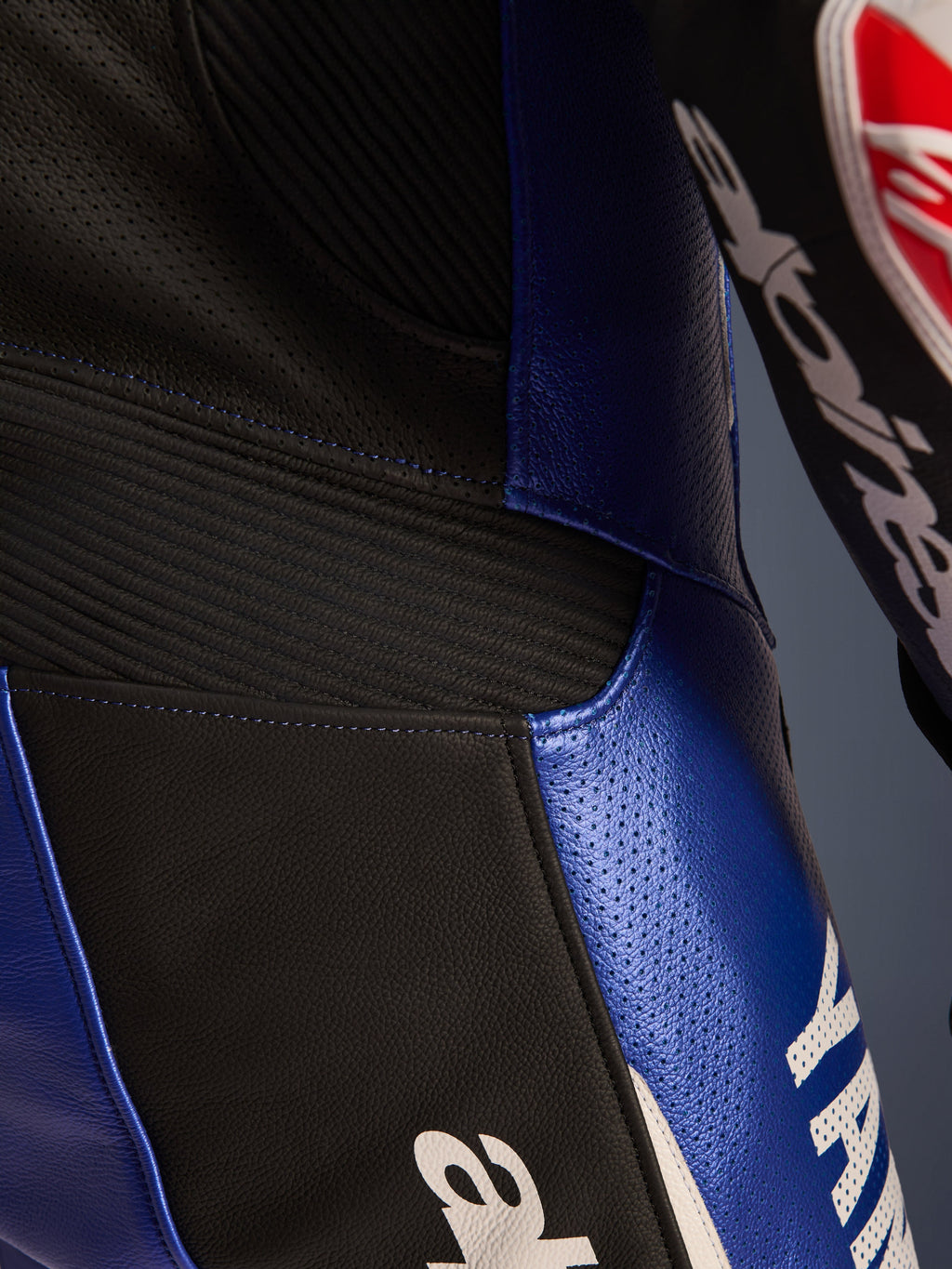 Nærbillede af detajler på Alpinesta rs Yamaha G P-R7 1-delt l æderdragt, metallic blå og sor t, med perfo rere de læ derpaneler ti l ventilation, sorte harmoni k a-st ræk inds atser for fle ksibilitet og markant hvid Ya ma h a-br anding, d er f re mhæv er konstruktion en i bovint l æder af racing-kva litet.