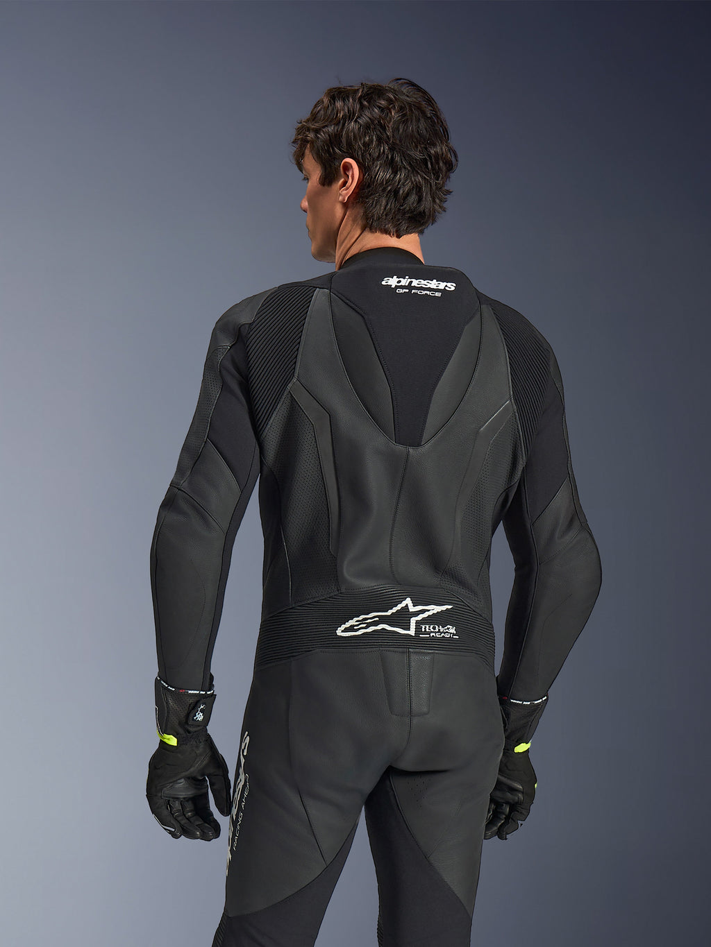 Alpinestars GP Force V2 læderdragt, 1-piece, sort, set bagfra med perforeret læder og harmonika-strækpaneler, hvidt Alpinestars- og Tech-Air Ready-branding på øverste og nederste del af ryggen, båret med sorte handsker, højtydende motorcykel-racerudstyr