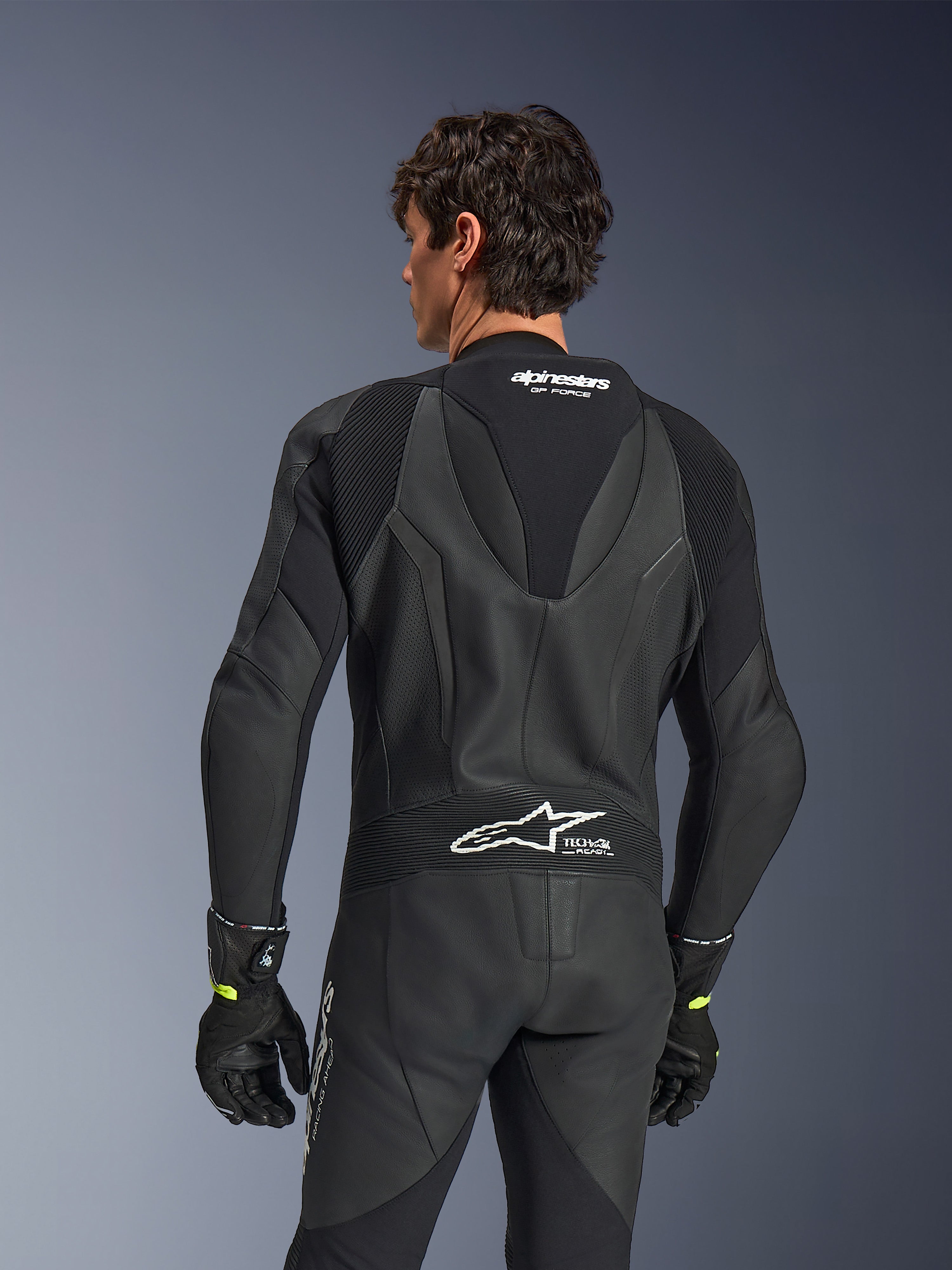 Alpinestars GP Force V2 læderdragt, 1-piece, sort, set bagfra med perforeret læder og harmonika-strækpaneler, hvidt Alpinestars- og Tech-Air Ready-branding på øverste og nederste del af ryggen, båret med sorte handsker, højtydende motorcykel-racerudstyr