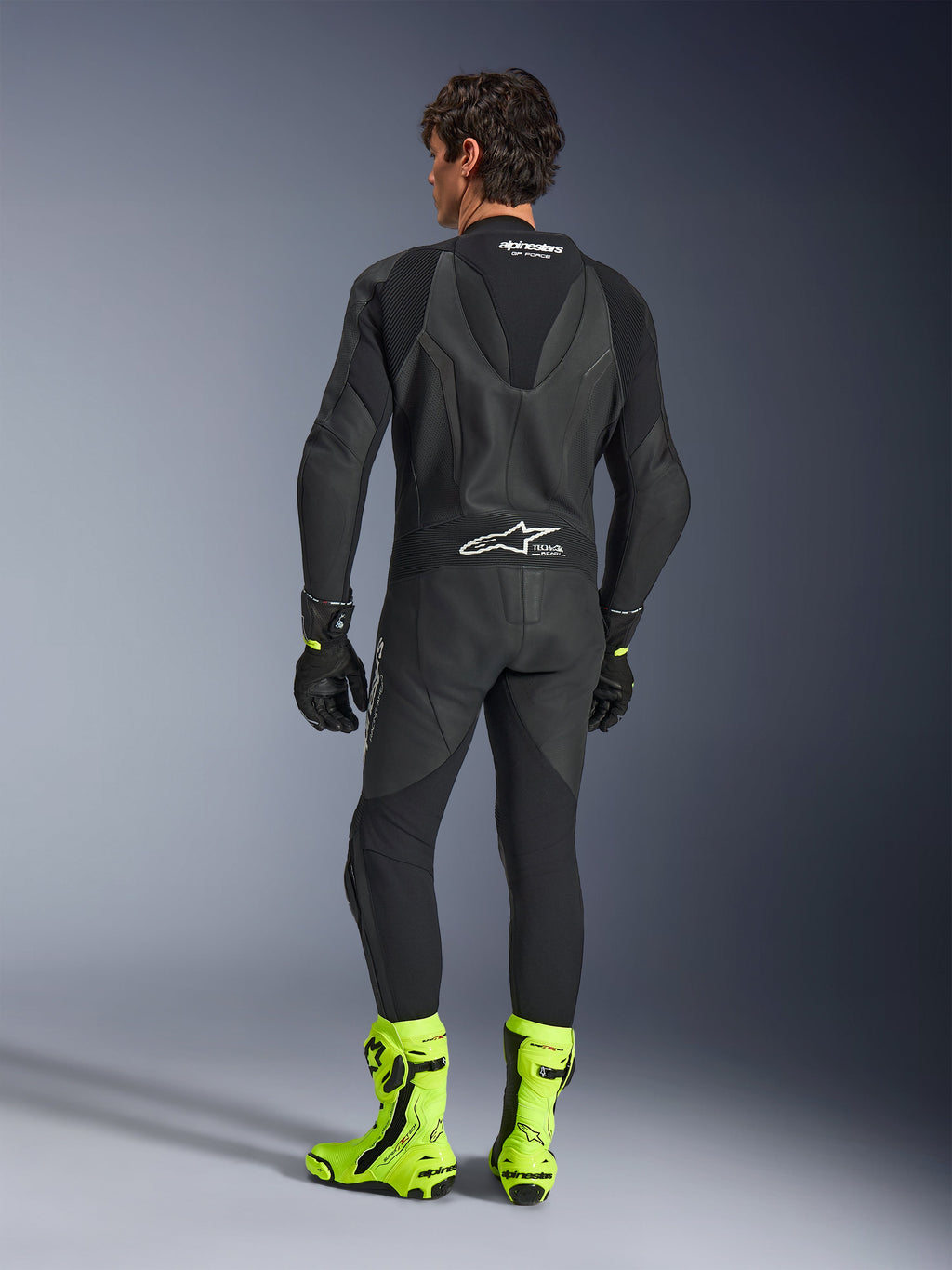 Alpinestars GP Force V2 læderdragt - 1-piece, sort, set bagfra båret af en model, med en aerodynamisk speed hump og Tech-Air Ready-branding, stylet med sorte handsker og klare fluorescerende gule racestøvler, professionelt baneudstyr