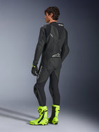 Alpinestars GP Force V2 læderdragt, 1-piece sort motorcykelracerdragt, set bagfra med aerodynamisk speed hump og perforeret læder, båret med neongule racerstøvler og sorte handsker, professionelt baneklart udstyr