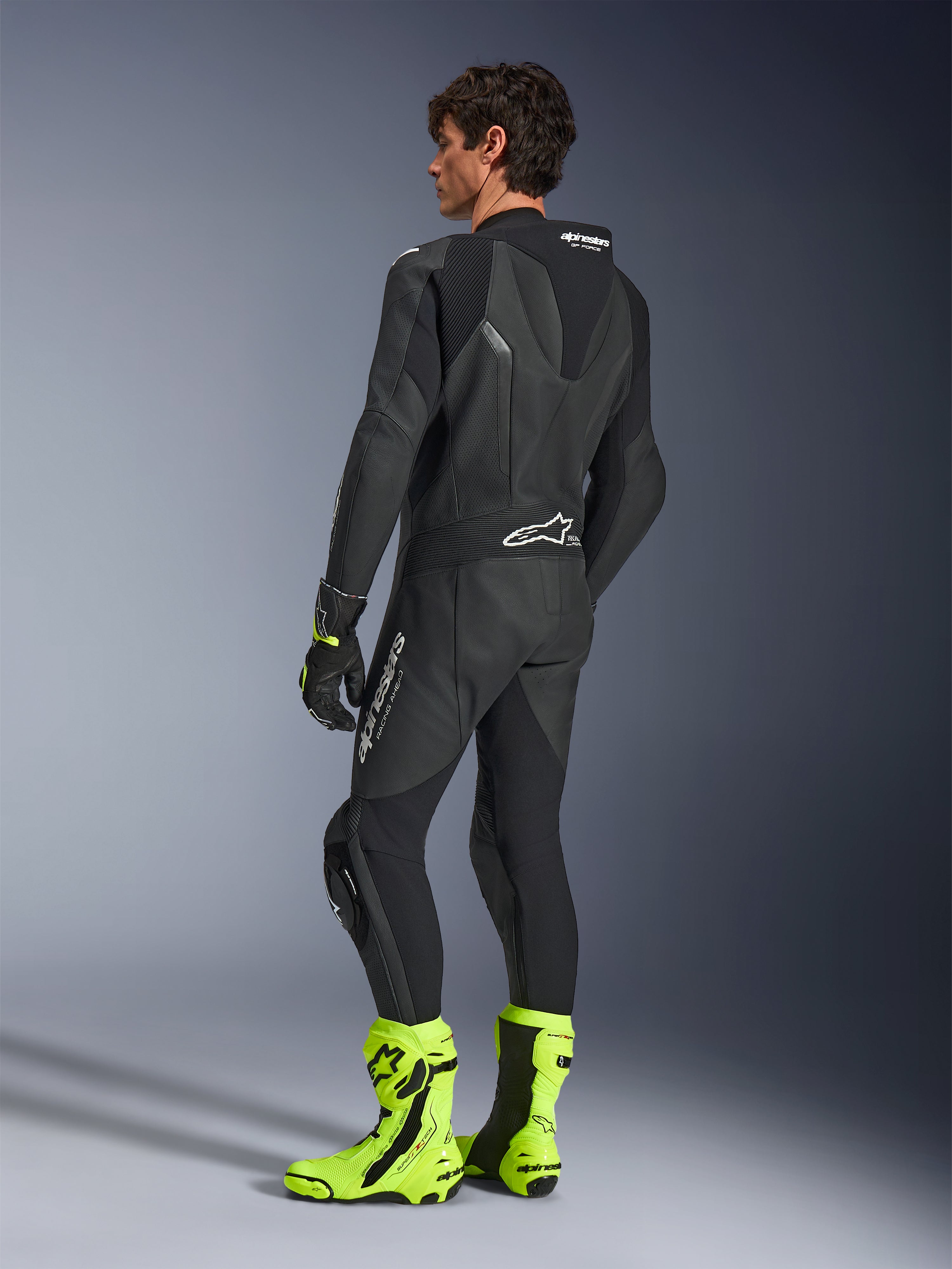 Alpinestars GP Force V2 læderdragt, 1-piece sort motorcykelracerdragt, set bagfra med aerodynamisk speed hump og perforeret læder, båret med neongule racerstøvler og sorte handsker, professionelt baneklart udstyr
