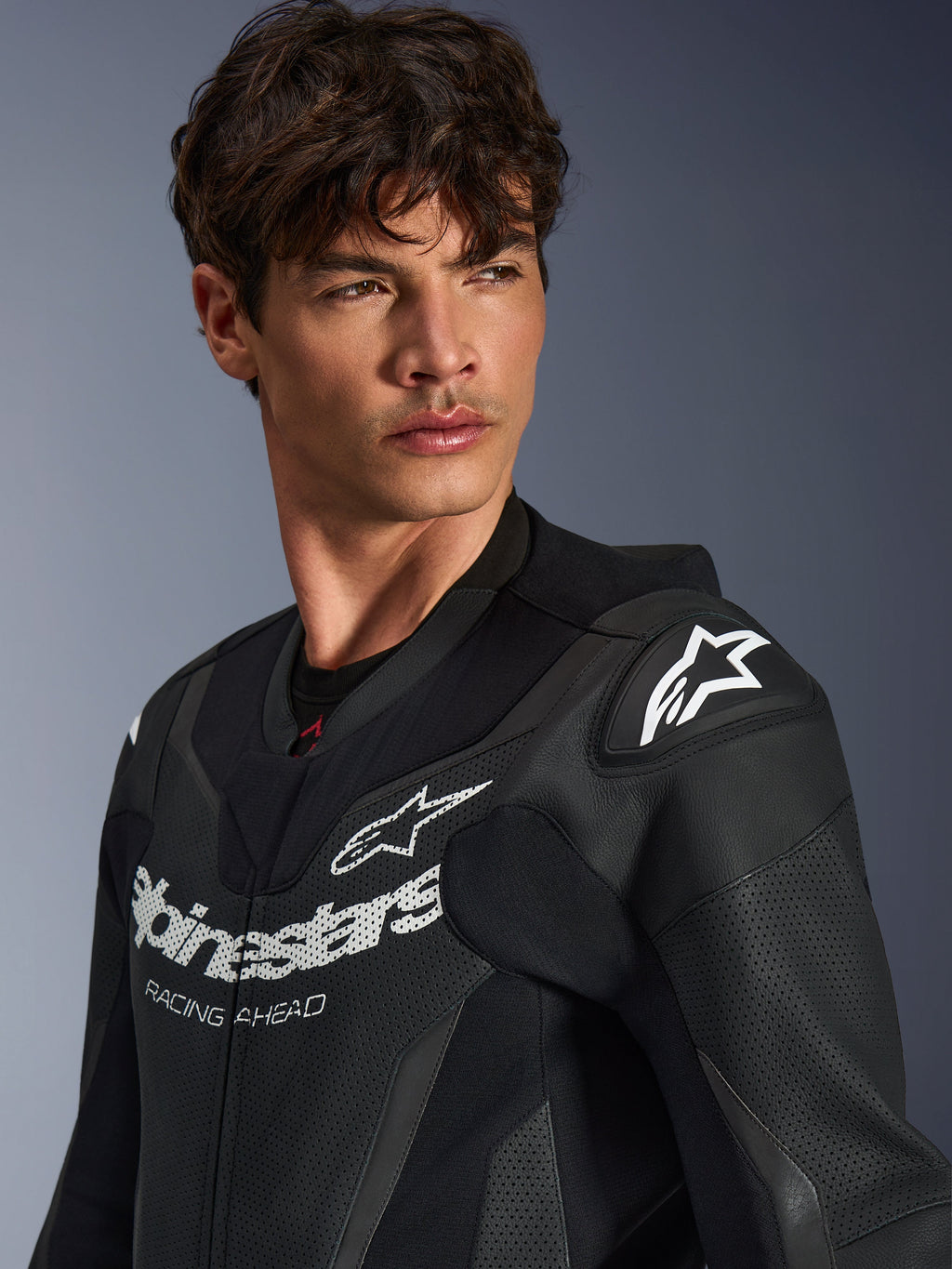 Nærbillede af en mand iført Alpinestars GP Force V2 læderdragt i sort, med perforeret læderkonstruktion, Alpinestars-logo med 'RACING AHEAD' branding på brystet og hvidt stjernelogo på skulderbeskytterne.