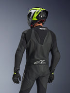 Alpinestars GP Force V2 Læderdragt 1-piece, sort, set bagfra med aerodynamisk speed hump og perforeret læderkonstruktion, båret med en sort og neongul racerhjelm og sorte handsker, med harmonika-strækpaneler og hvid Tech-Air Ready-branding.
