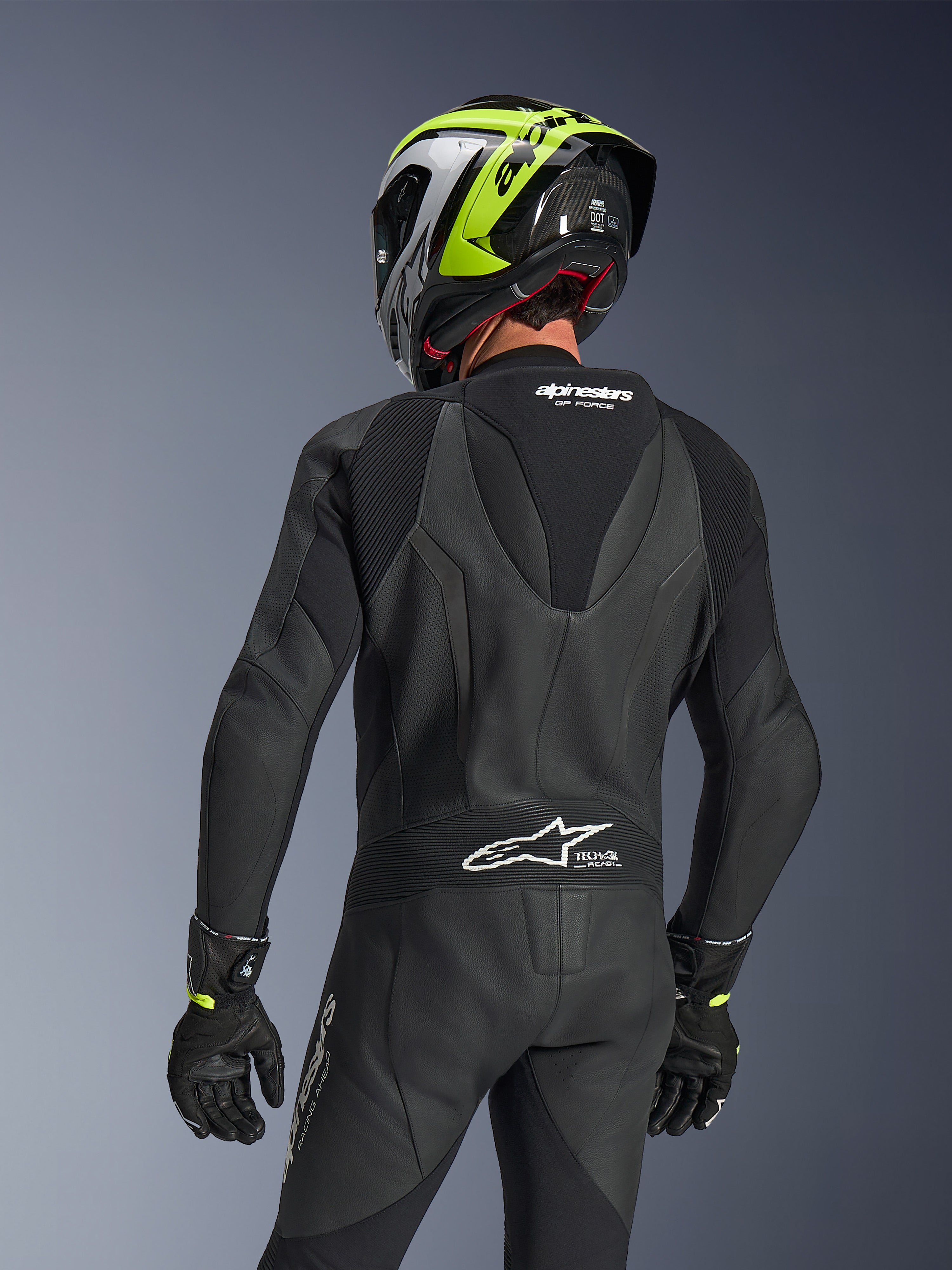 Alpinestars GP Force V2 Læderdragt 1-piece, sort, set bagfra med aerodynamisk speed hump og perforeret læderkonstruktion, båret med en sort og neongul racerhjelm og sorte handsker, med harmonika-strækpaneler og hvid Tech-Air Ready-branding.