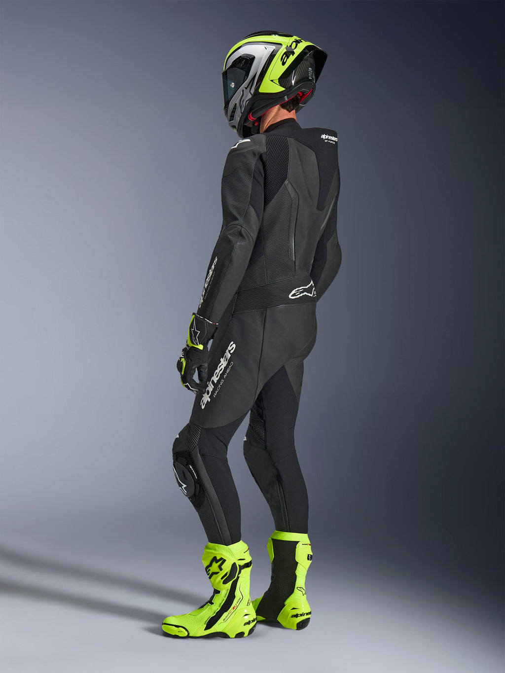 Motorcykelrytter iført Alpinestars GP Force V2 læderdragt 1-piece i sort, set bagfra med hvid branding på låret og lænden, aerodynamisk pukkel, kombineret med en neongul og sort hjelm, matchende racerhandsker og støvler, professionelt udstyr til bane- og sportskørsel.