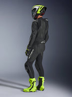Motorcykelrytter iført Alpinestars GP Force V2 læderdragt 1-piece i sort, set bagfra med hvid branding på låret og lænden, aerodynamisk pukkel, kombineret med en neongul og sort hjelm, matchende racerhandsker og støvler, professionelt udstyr til bane- og sportskørsel.