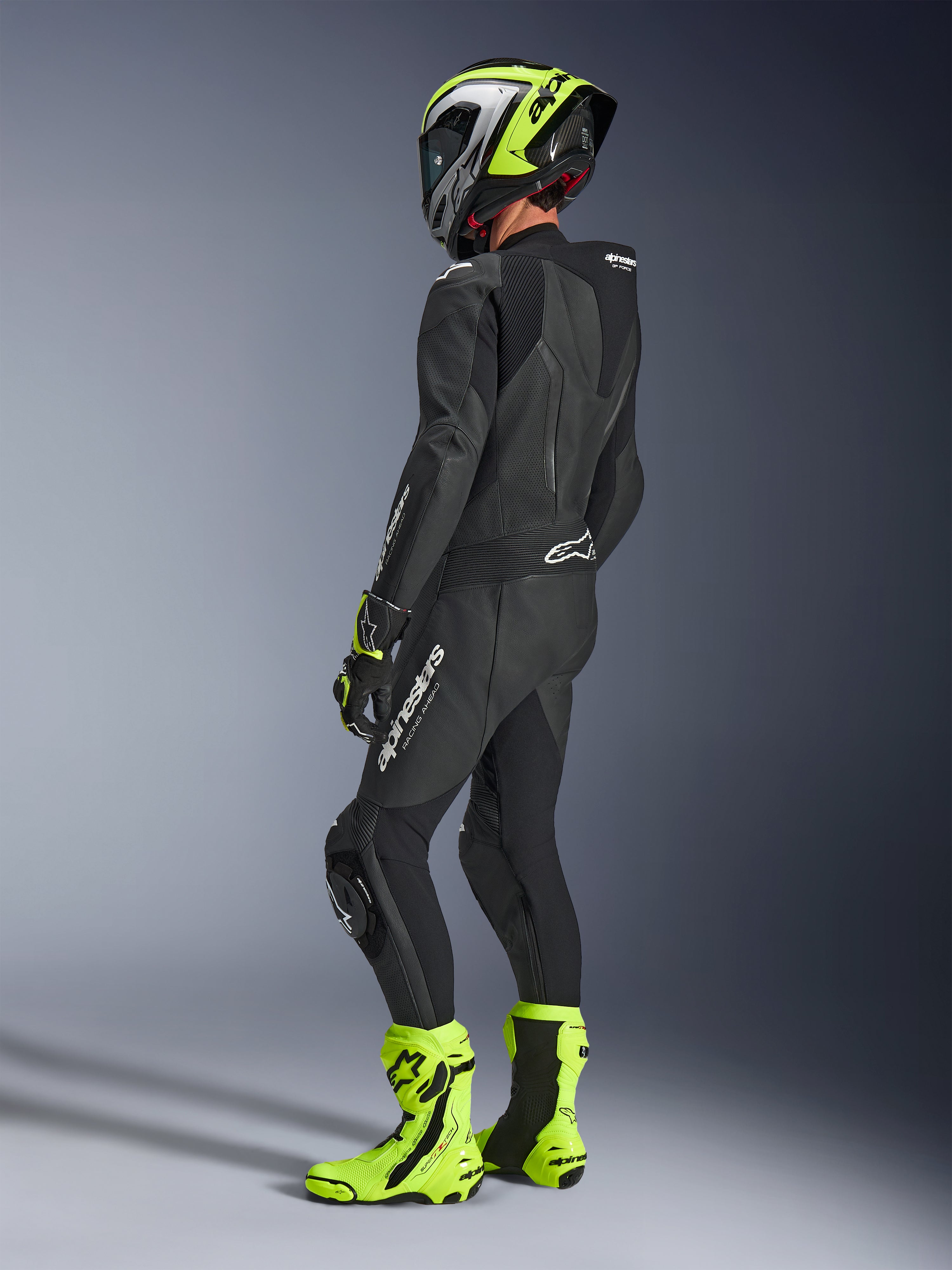 Motorcykelrytter iført Alpinestars GP Force V2 læderdragt 1-piece i sort, set bagfra med hvid branding på låret og lænden, aerodynamisk pukkel, kombineret med en neongul og sort hjelm, matchende racerhandsker og støvler, professionelt udstyr til bane- og sportskørsel.