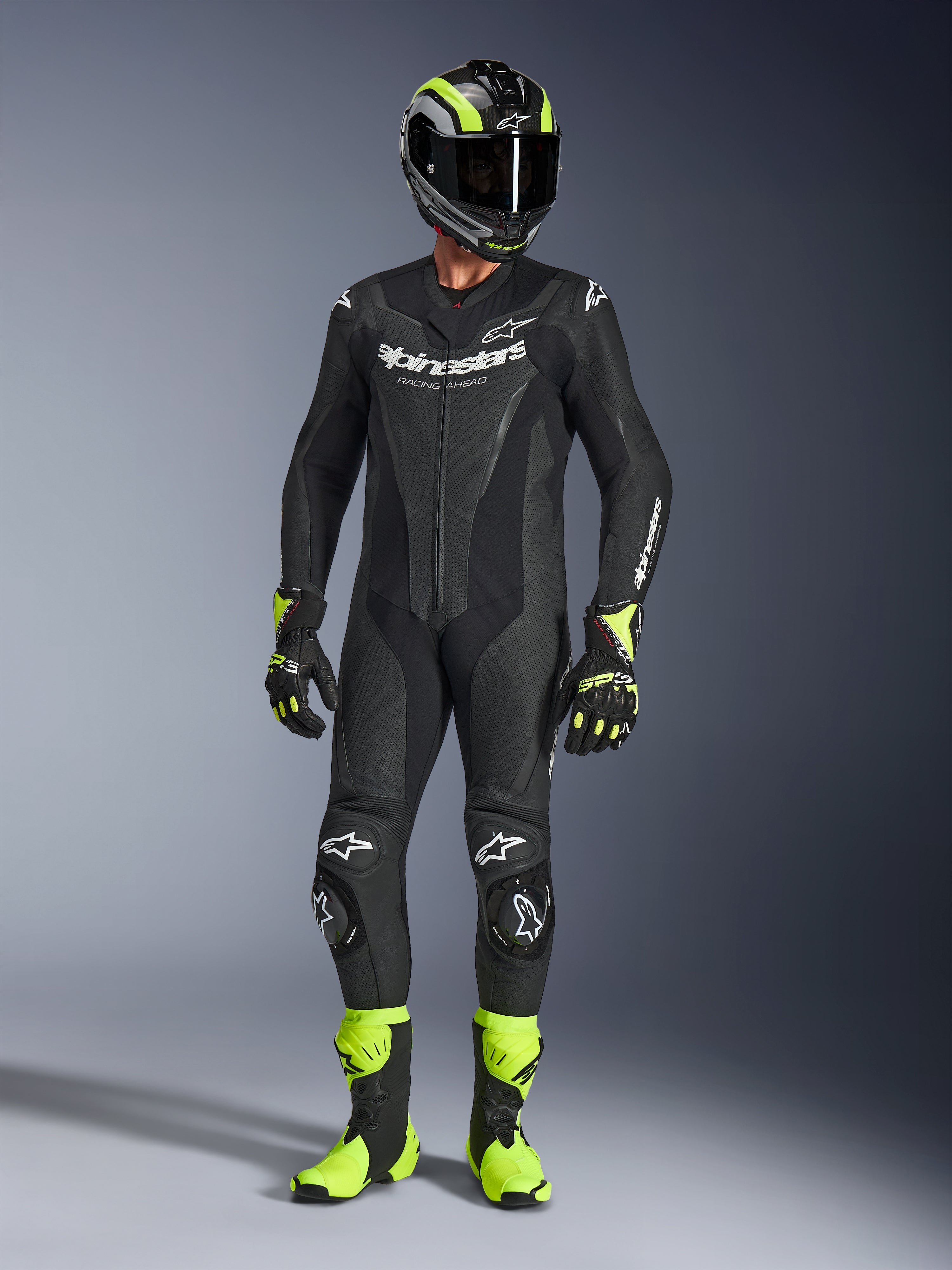 Motorcykelrytter iført Alpinestars GP Force V2 læderdragt i sort, 1-delt læderdragt med hvid branding, kombineret med en sort og neongul racinghjelm, handsker og støvler, professionelt banekørselsudstyr på en grå studiebaggrund