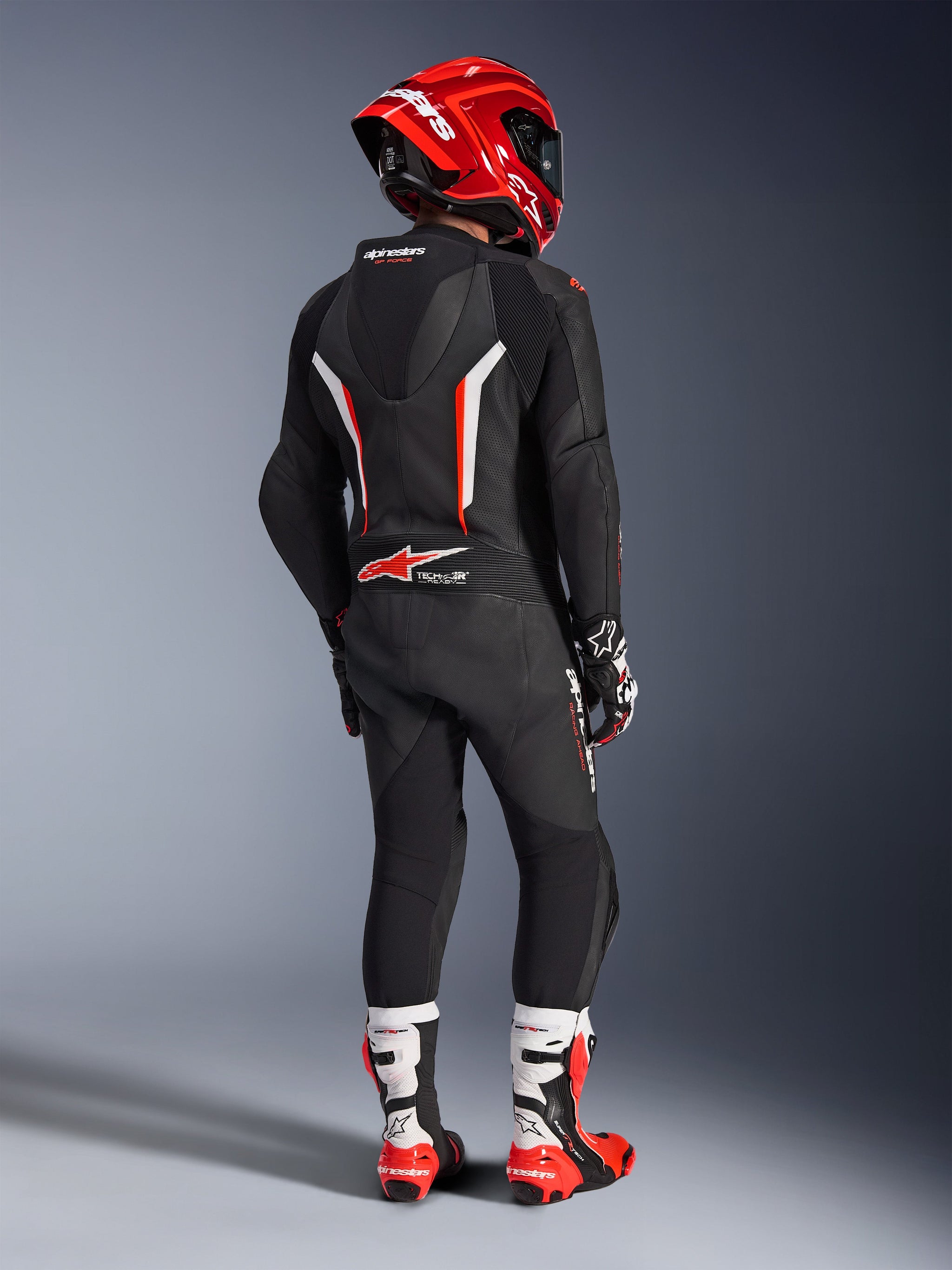 Alpinestars GP Force V2 1-delt læderdragt, kører set bagfra i sort, hvid og rød fluo, med aerodynamisk pukkel og perforerede læderpaneler, rød Alpinestars hjelm, racerhandsker og rød-hvide motorcykelstøvler, professionelt racerudstyr til banekørsel