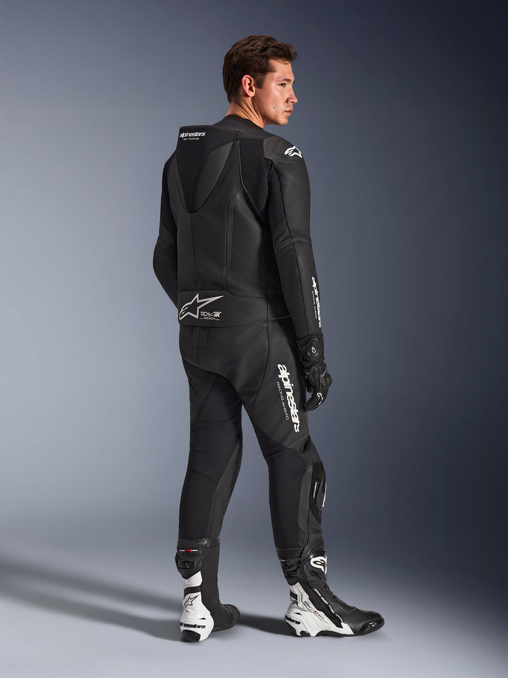 Mandlig model iført Alpinestars GP Force V2 2-delt læderdragt i sort med hvide logoer, set bagfra i trekvart profil, der viser den aerodynamiske speed hump og Tech-Air Ready-branding, kombineret med sort-hvide racingstøvler og handsker, professionelt motorcykeludstyr til sport og banekørsel.