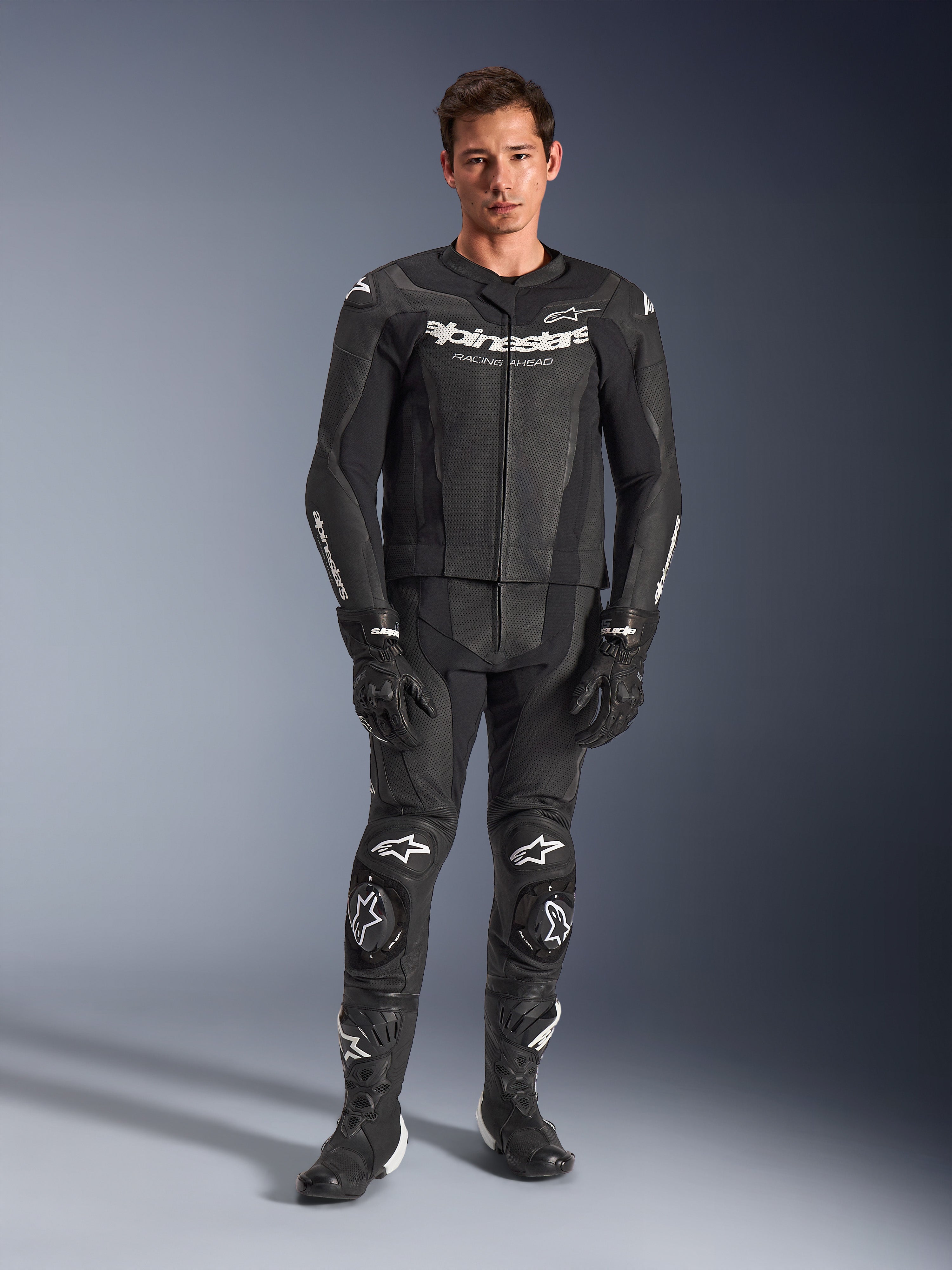 Alpinestars GP Force V2 todelt læderdragt, sort med hvide logoer, fuld motorcykelracingdragt med premium læderkonstruktion, forstærkede knæslidere og perforerede paneler, båret med sorte handsker og støvler, studieoptagelse i fuld længde