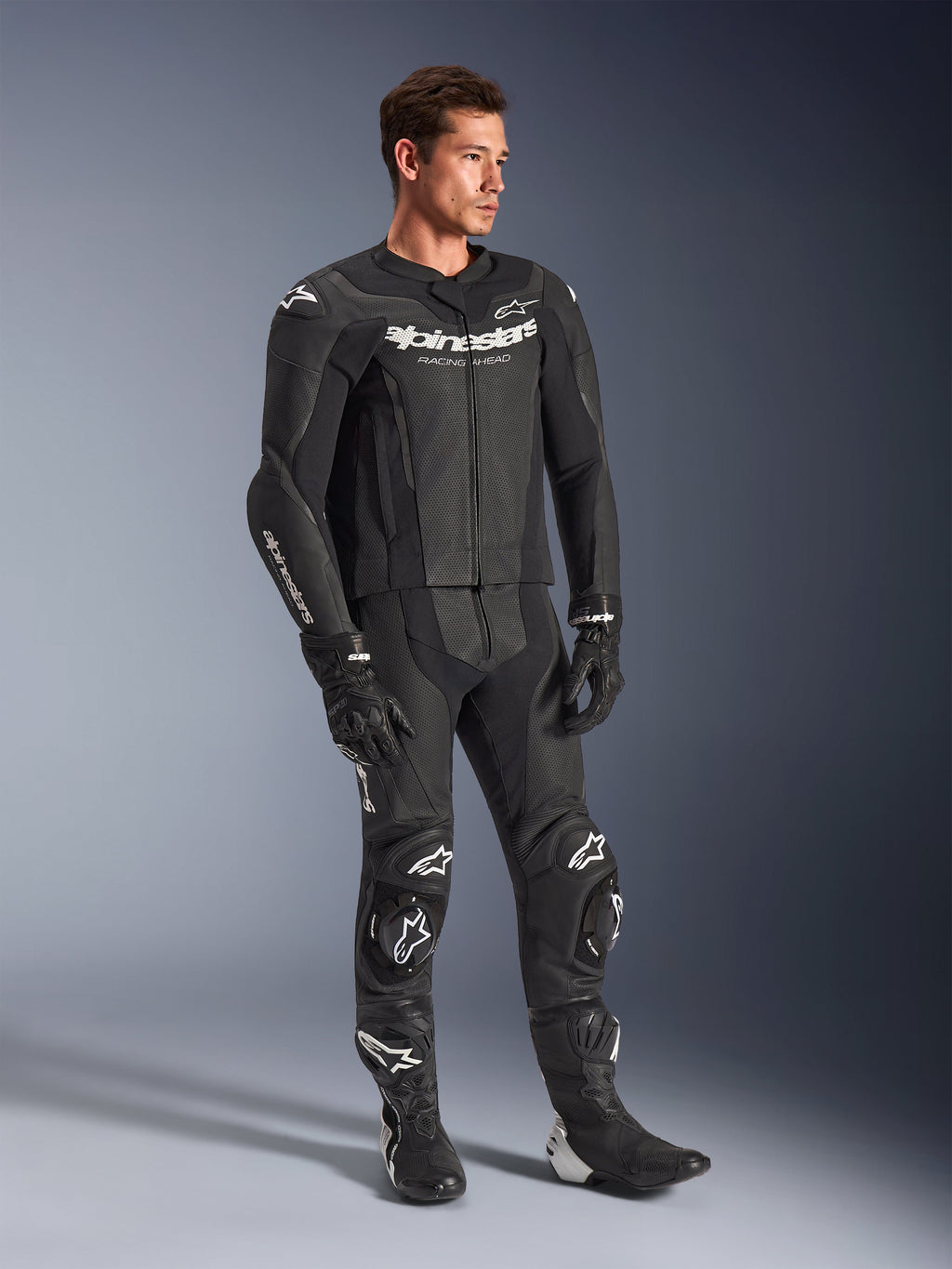 Alpinestars GP Force V2 2-delt læderdragt, sort, båret af en mandlig model med matchende sorte racerhandsker og støvler, med hvidt Alpinestars-logo, perforerede læderpaneler for ventilation og integrerede knæslidere, professionelt motorcykelracerudstyr