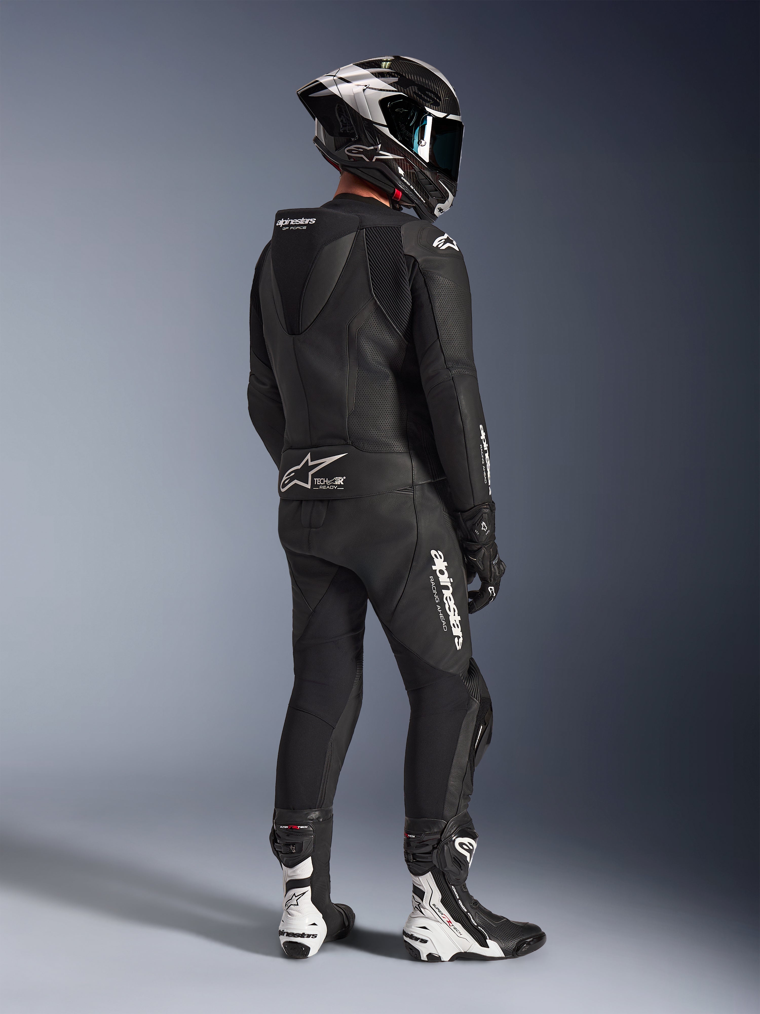 Alpinestars GP Force V2 2-delt læderdragt, sort, motorcykelrytter set bagfra i fuldt racerudstyr, med aerodynamisk fartpukkel, hvid branding og perforeret læder, kombineret med en kulfiberhjelm og sort-hvide racerstøvler