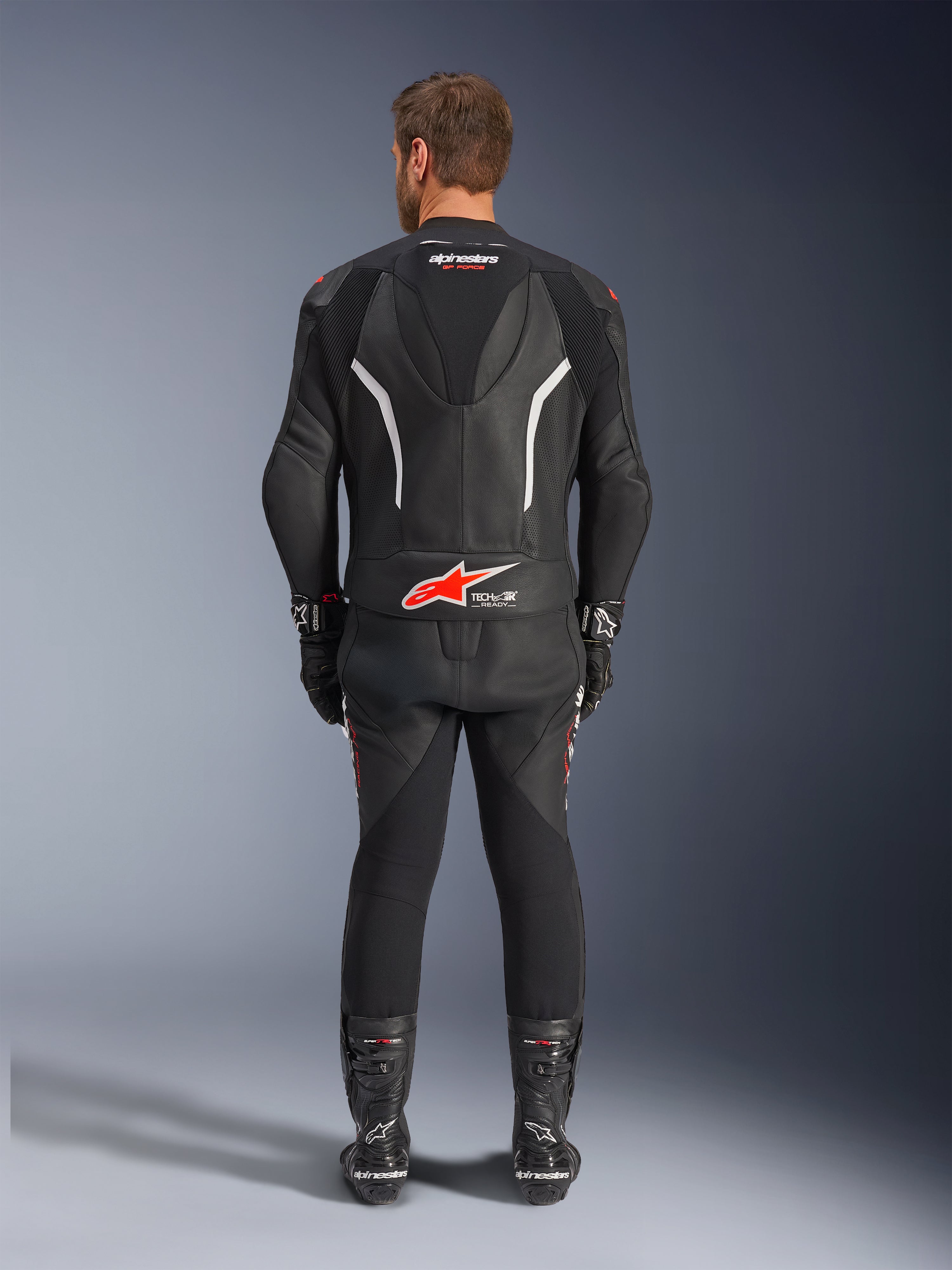 Rear view of a rider wearing the Alpinestars GP Force V2 2-Piece Leather Suit, Black with white and red accents, featuring prominent Alpinestars and Tech-Air Ready branding on the back, styled with matching black racing gloves and boots.