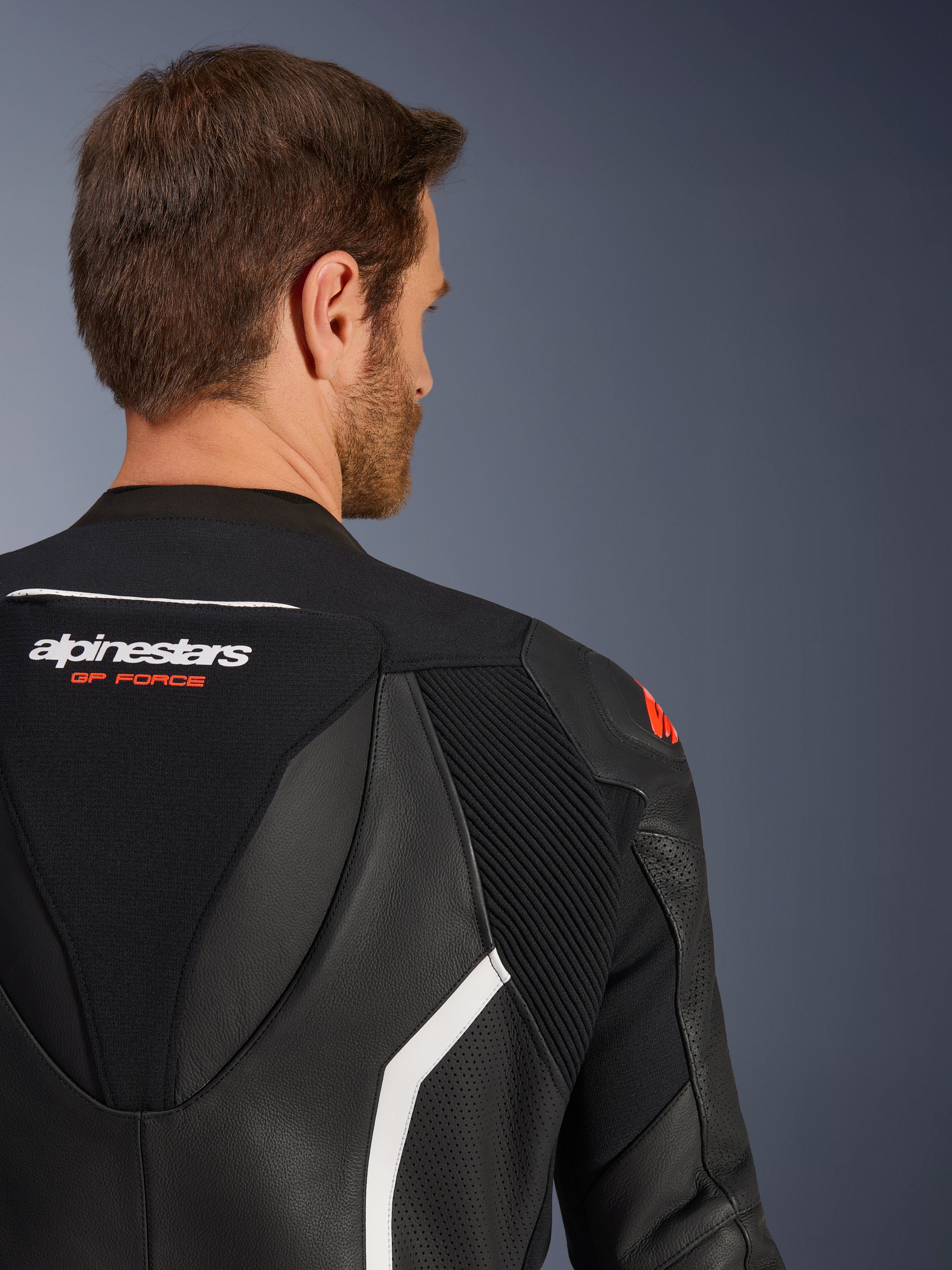 GP Force V2 2-Piece Leather Suit, close-up of the upper back and shoulder on a male model, black leather with white and red accents, Alpinestars GP Force branding, showing perforated panels and accordion stretch zones for motorcycle racing.