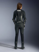 Female rider shown from the back wearing the Alpinestars Stella GP-R7 1-Piece Leather Suit, black leather with white branding, aerodynamic speed hump with GP-R7 logo, Tech-Air Ready designation, paired with black racing boots, professional motorcycle racing gear optimized for women